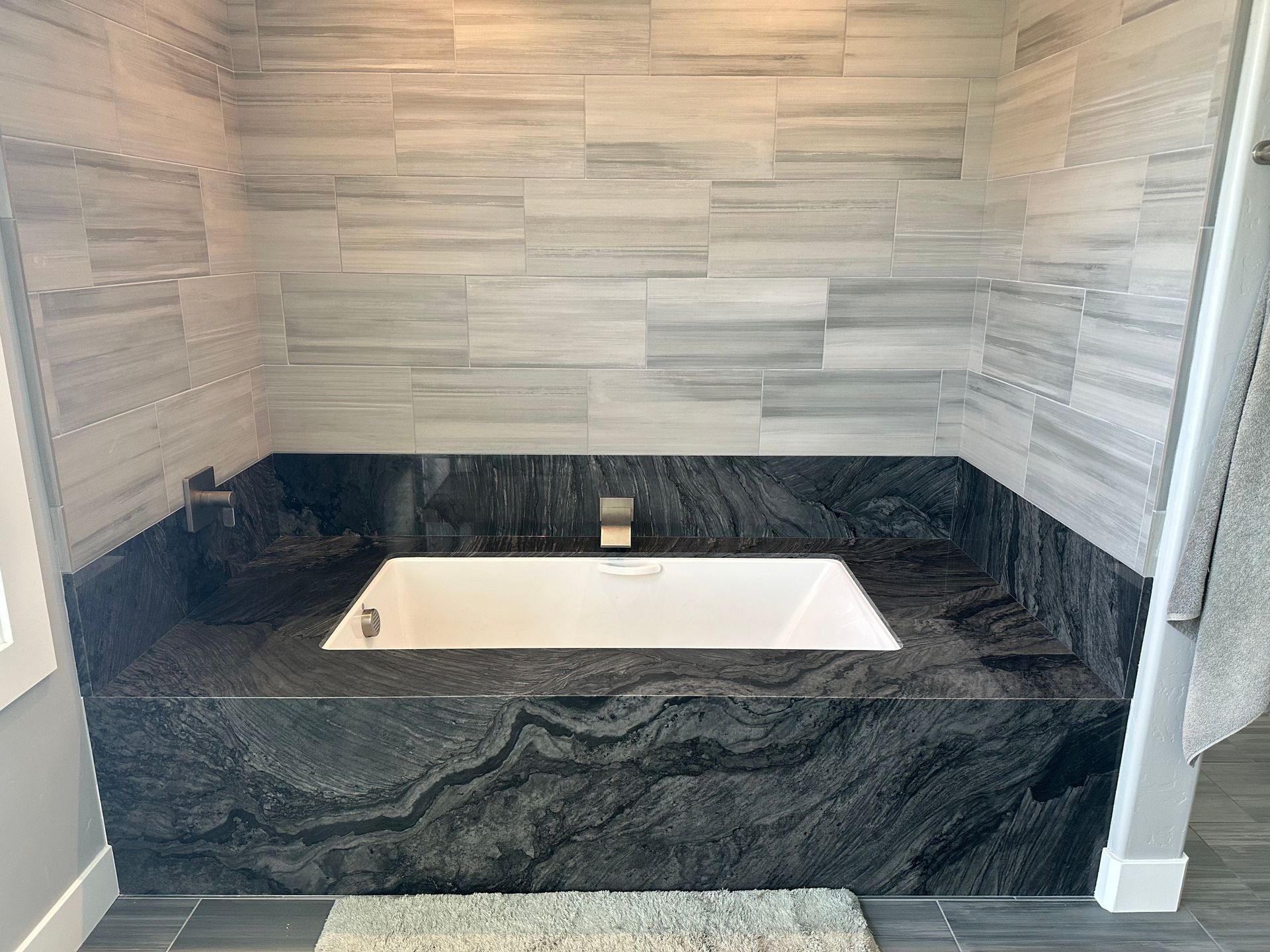 Bathroom with a white bathtub surrounded by dark granite and light gray tiled walls.