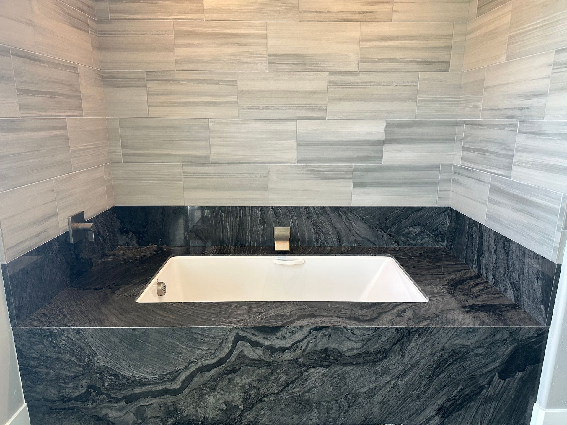 Bathroom with white rectangular tub, set in black granite countertop. Gray tile wall above.