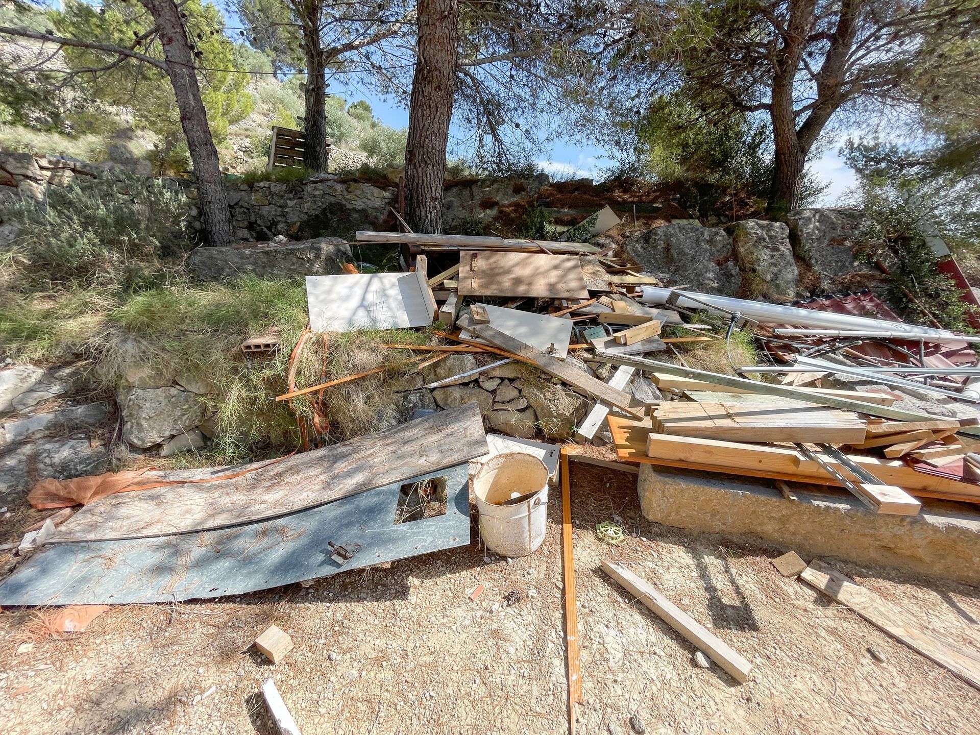 Pile of lumber, scrap wood, and debris in a wooded outdoor area with stone walls.