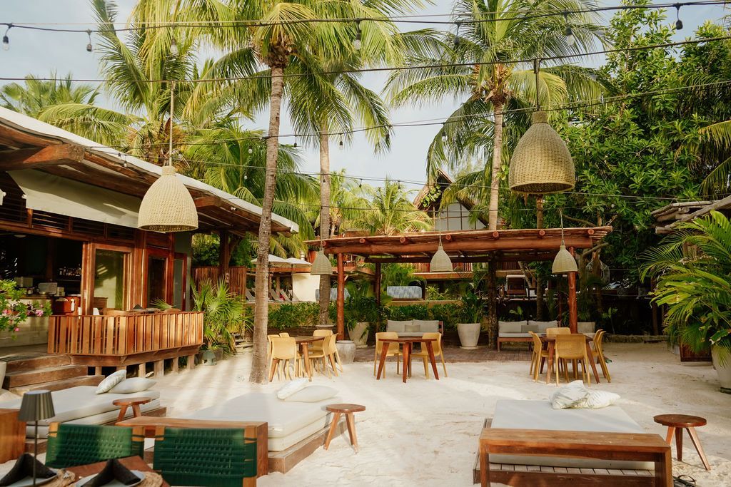 Outdoor restaurant with sand floor, wooden structures, palm trees, and hanging woven lamps.