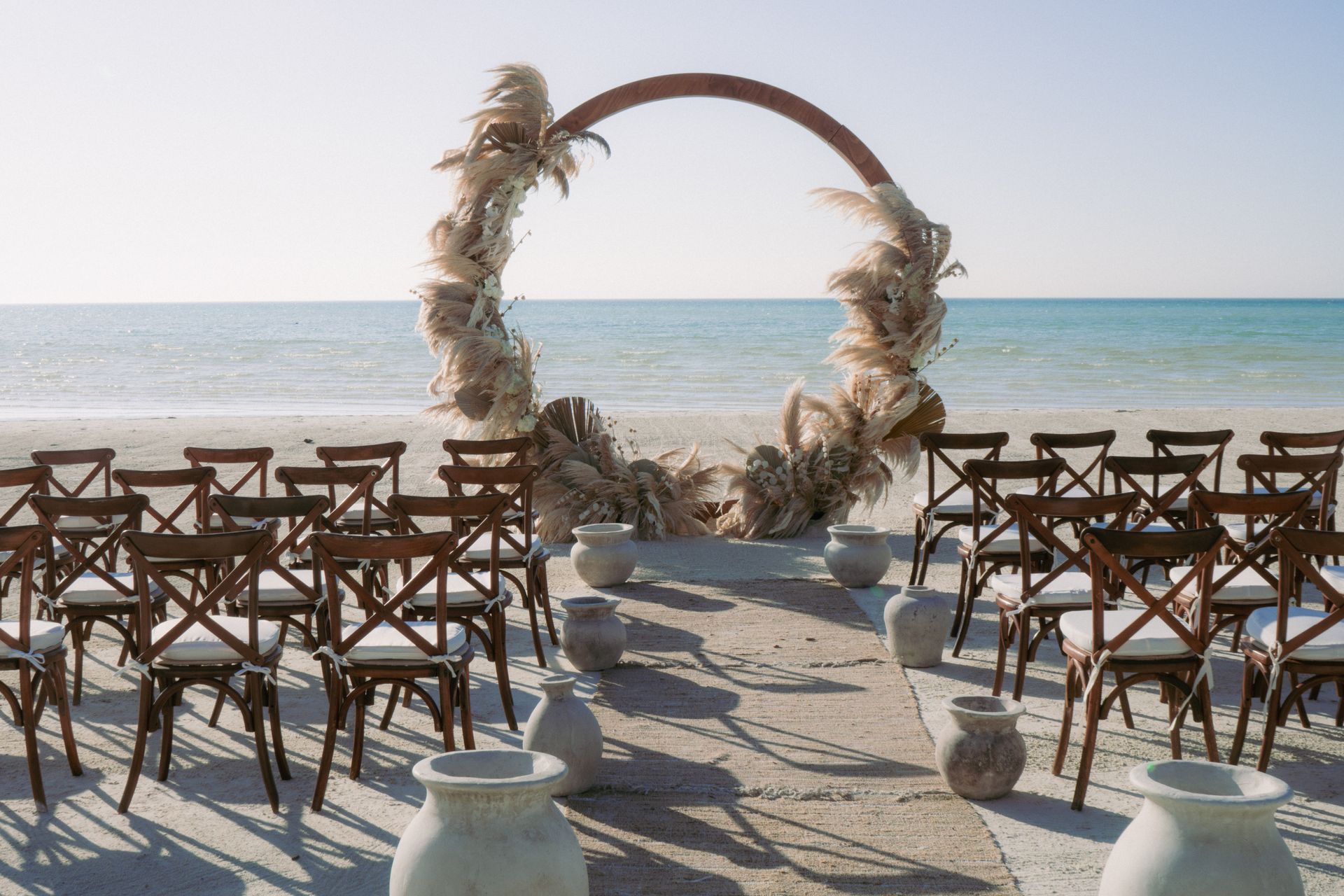 Beach wedding ceremony with arch, chairs, and vases.
