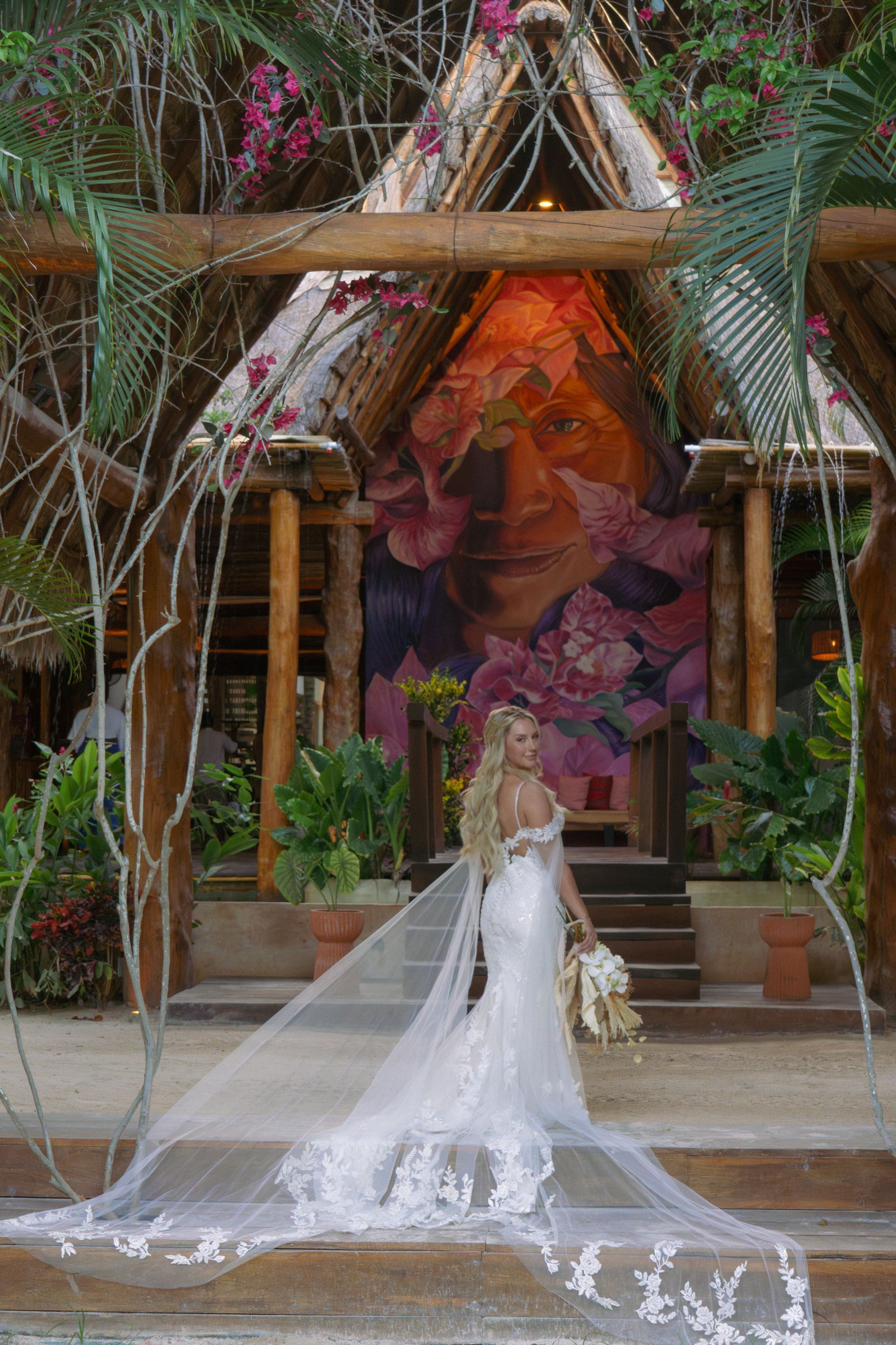 Bride in a white lace wedding dress, holding a bouquet, stands beneath a wooden arch with a painting as a backdrop.