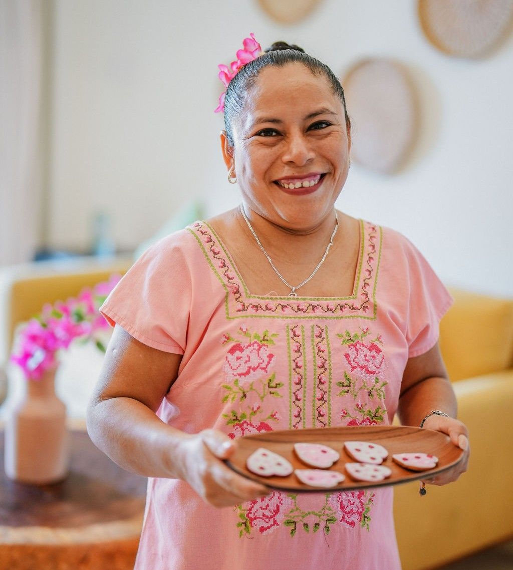 Woman holding tray of cookies, smiling, wearing pink embroidered dress, indoors.