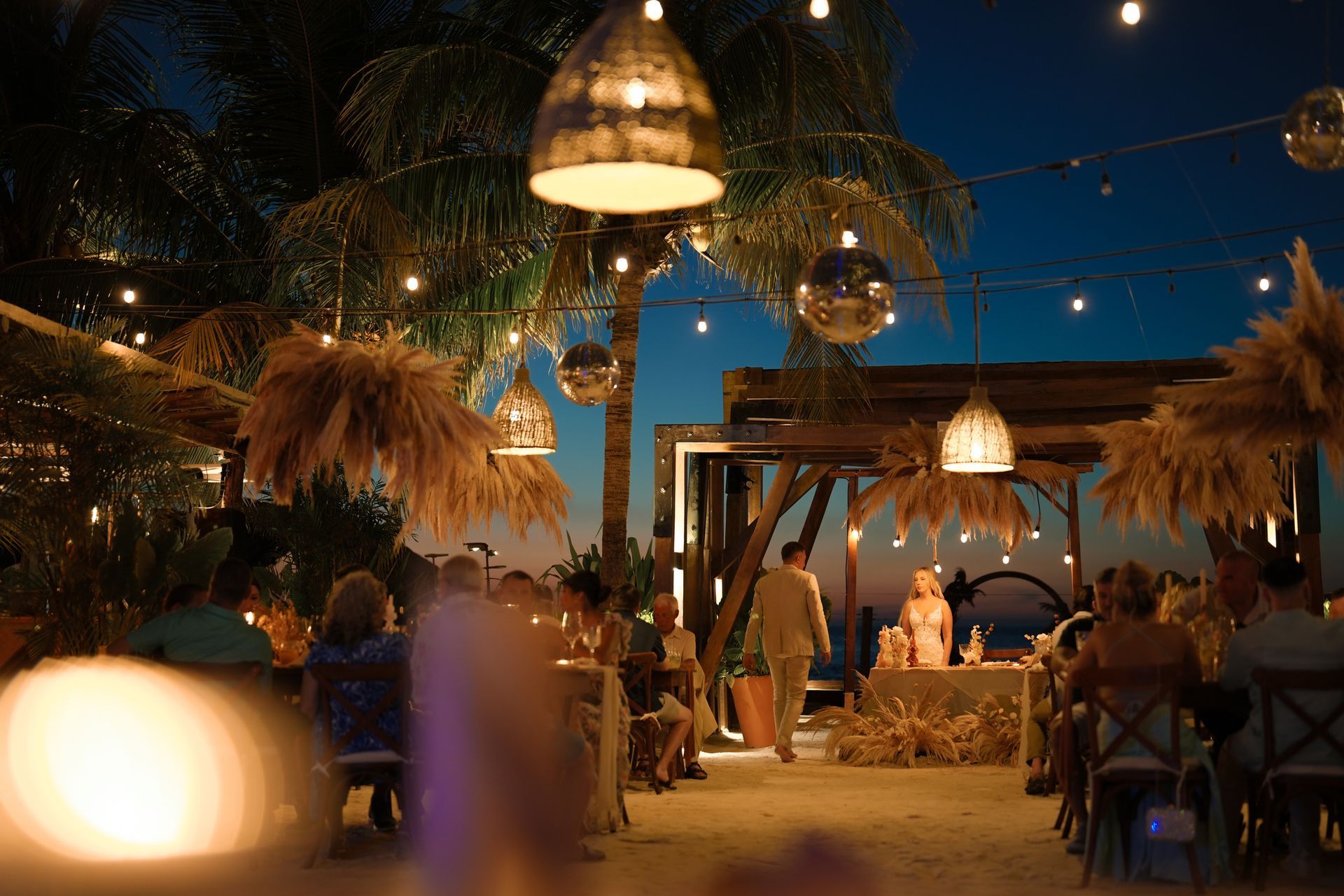 Beach wedding at dusk with string lights, disco balls, and guests seated. Couple stands at a decorated table.