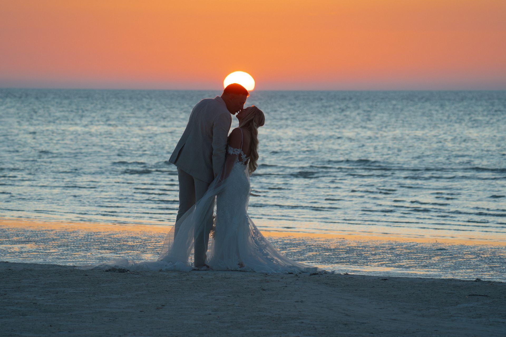 Couple kissing at sunset on a beach, woman in wedding dress, man in a suit, sun over the water.