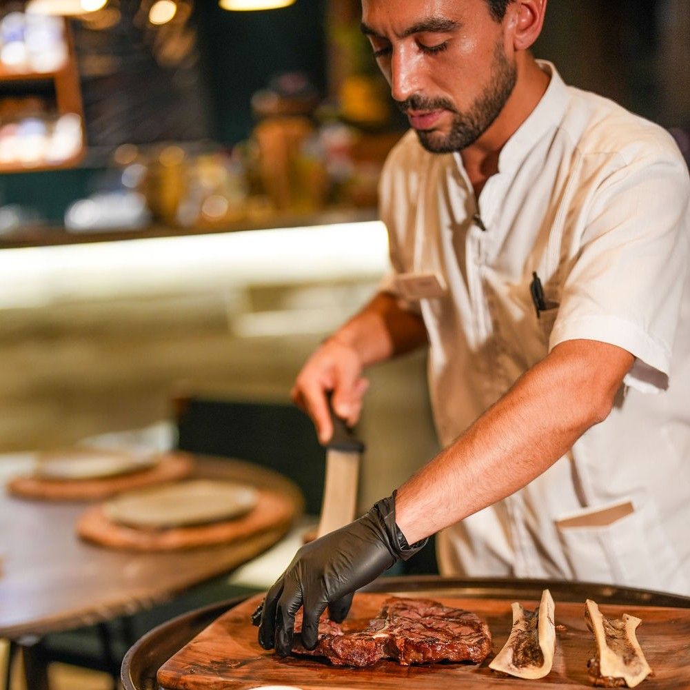 Chef carving a cooked steak on a wooden board; wearing black gloves and white chef coat, restaurant setting.