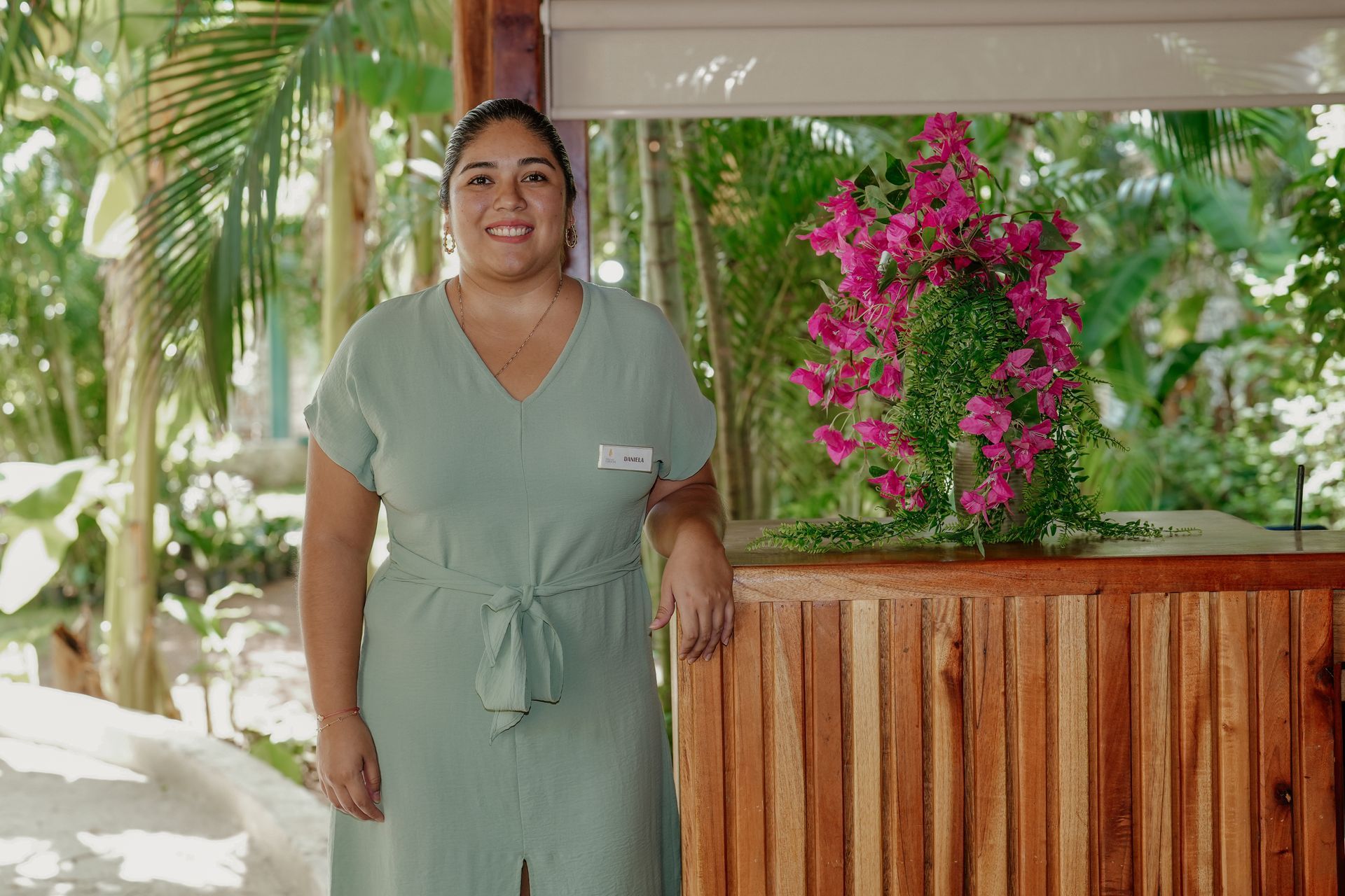Woman at a reception desk smiles; wearing a green uniform. Laptop and green foliage in the background.