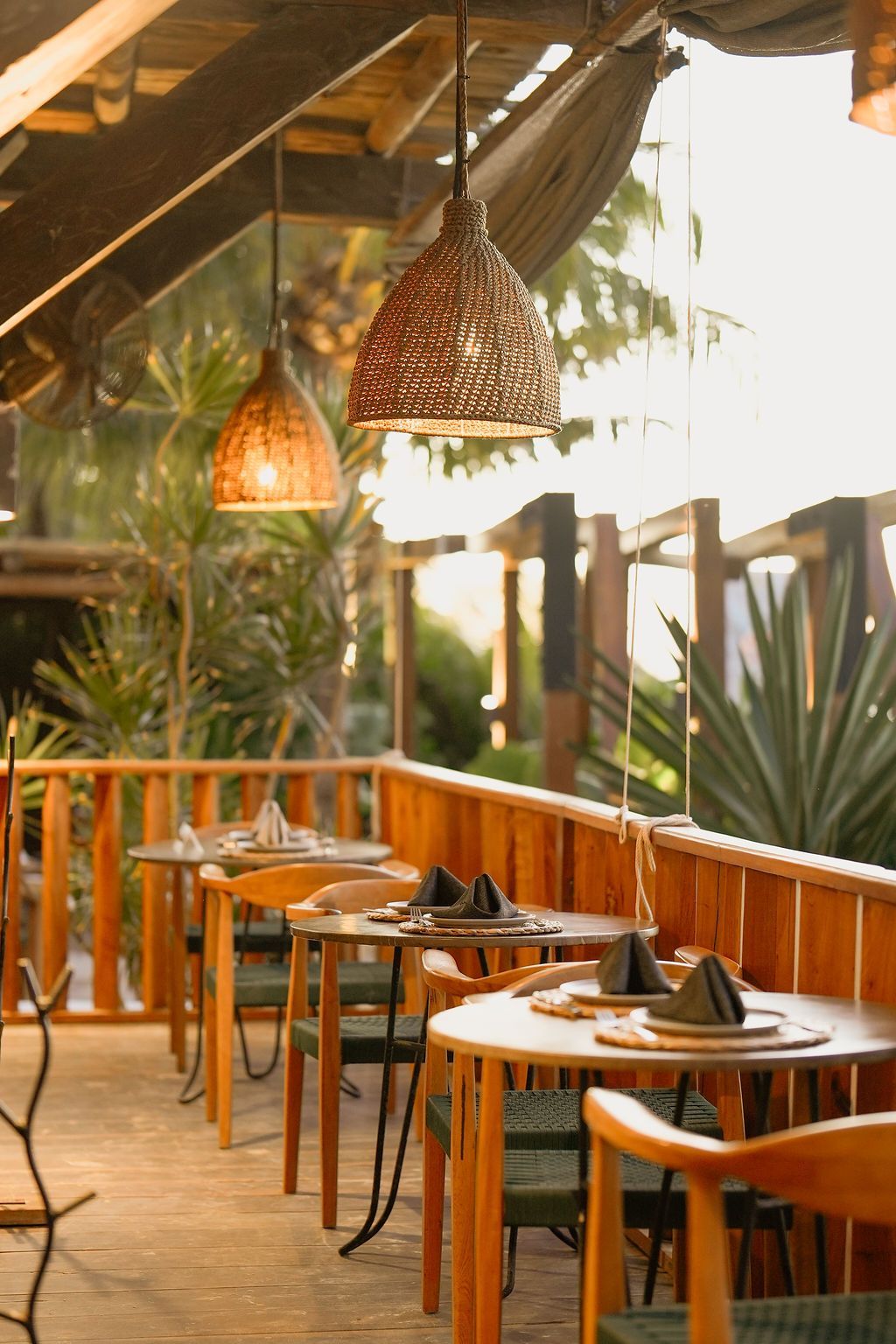 Outdoor restaurant patio with wood tables, chairs, and woven pendant lights.