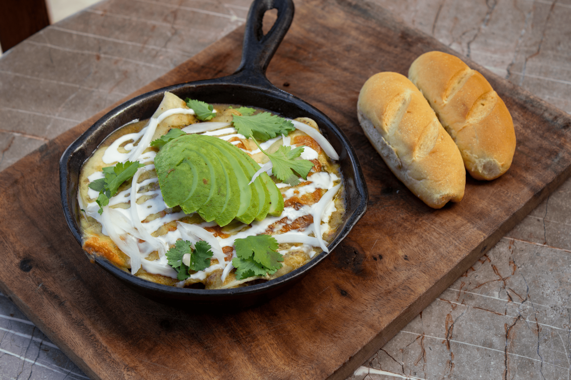 Enchiladas in a cast iron skillet, topped with avocado, cilantro, and crema. Served with bread rolls on a wood board.