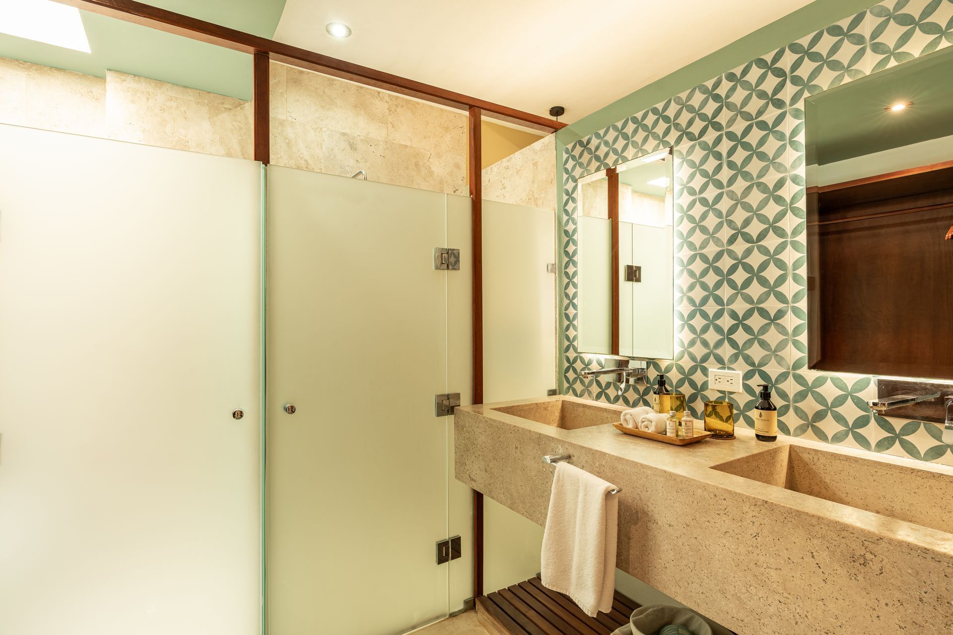 Bathroom with frosted glass shower, double sinks, patterned green tile, and wooden accents.