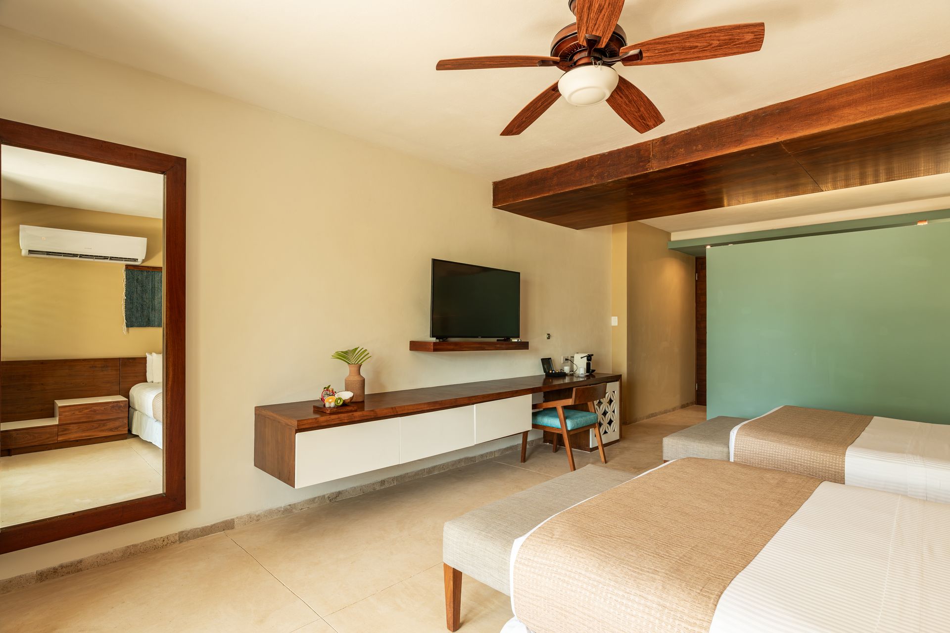 Hotel room with two beds, floating TV, and desk. Beige walls, wood accents, and a ceiling fan.