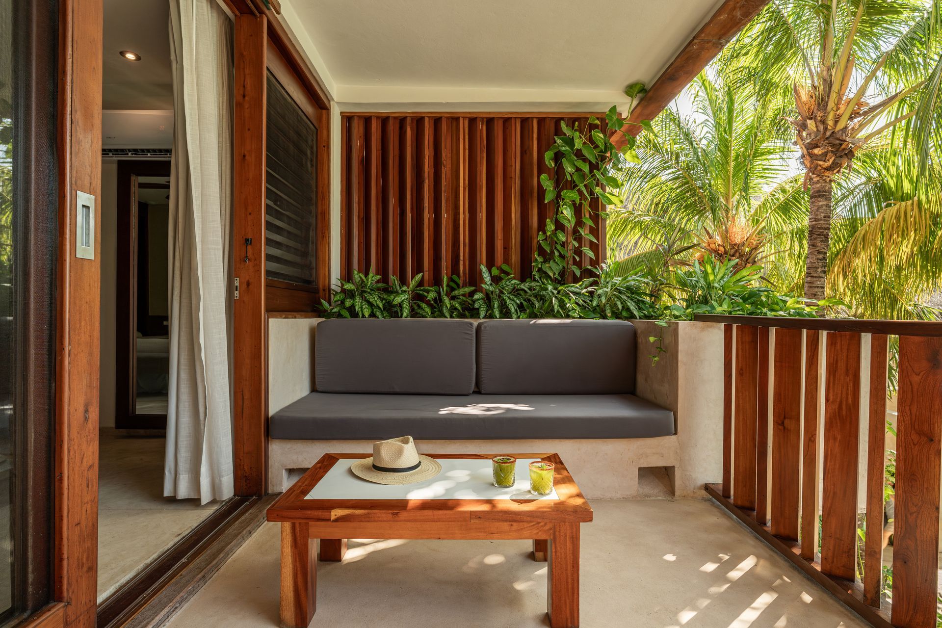 Balcony with wooden accents, gray cushion, small table, plants, and view of greenery.