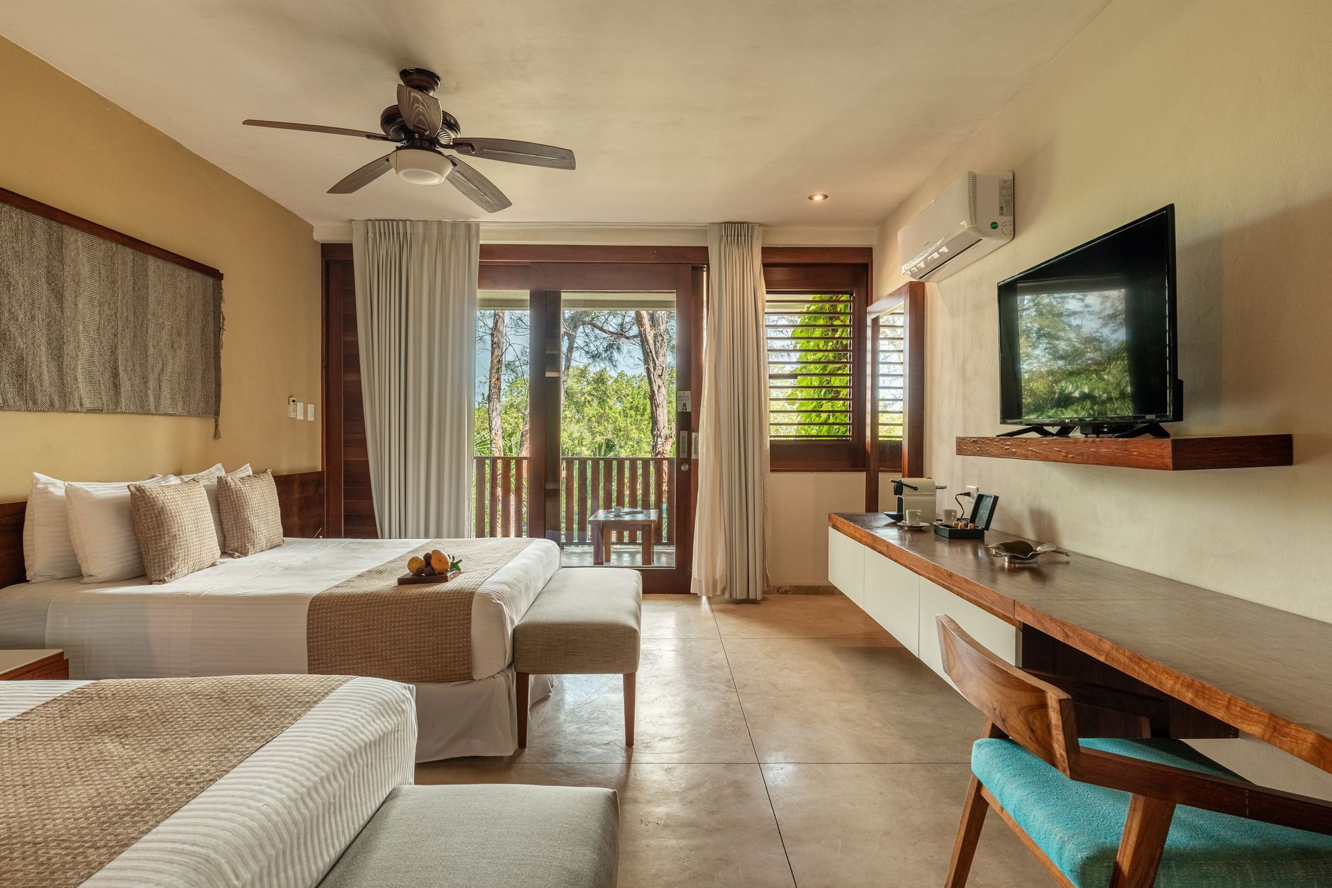 Hotel room with two beds, balcony access, TV, and a desk. Neutral colors, natural light.
