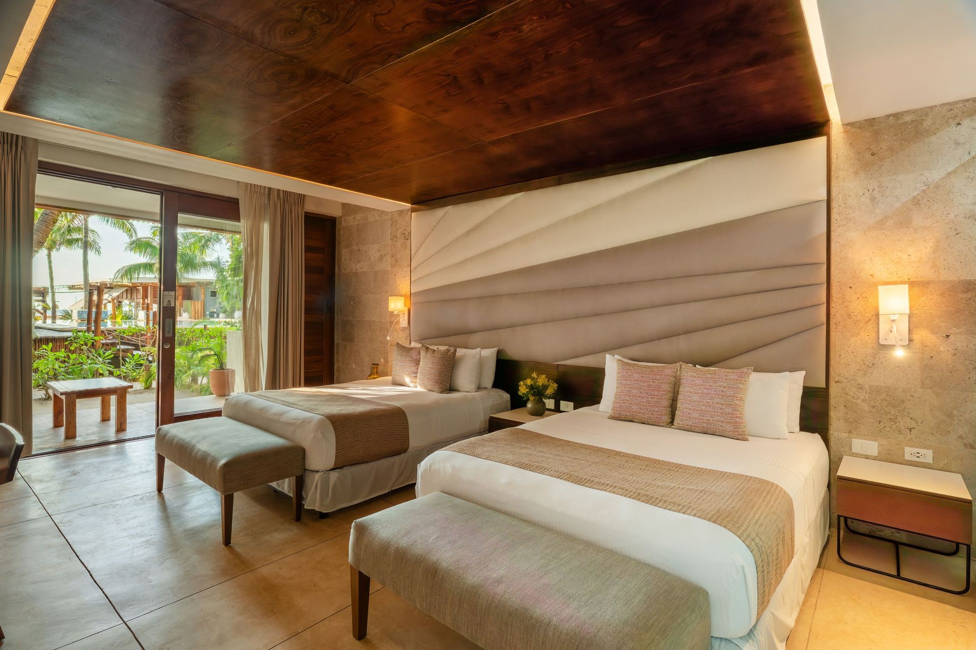Hotel room with two double beds, light wood ceiling, and a balcony overlooking greenery.