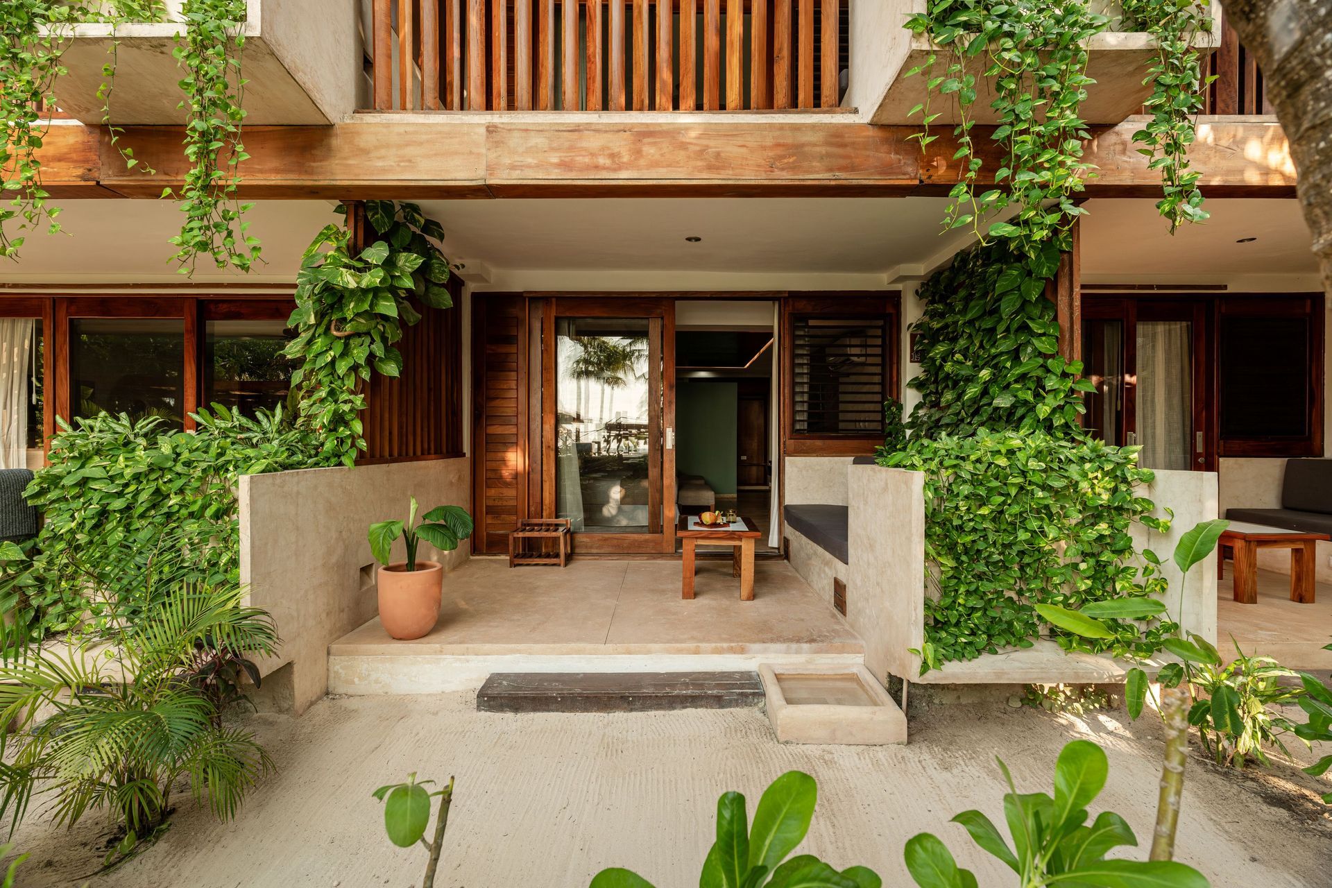 Exterior view of a beachside bungalow with wooden accents, lush greenery, and a sandy path.