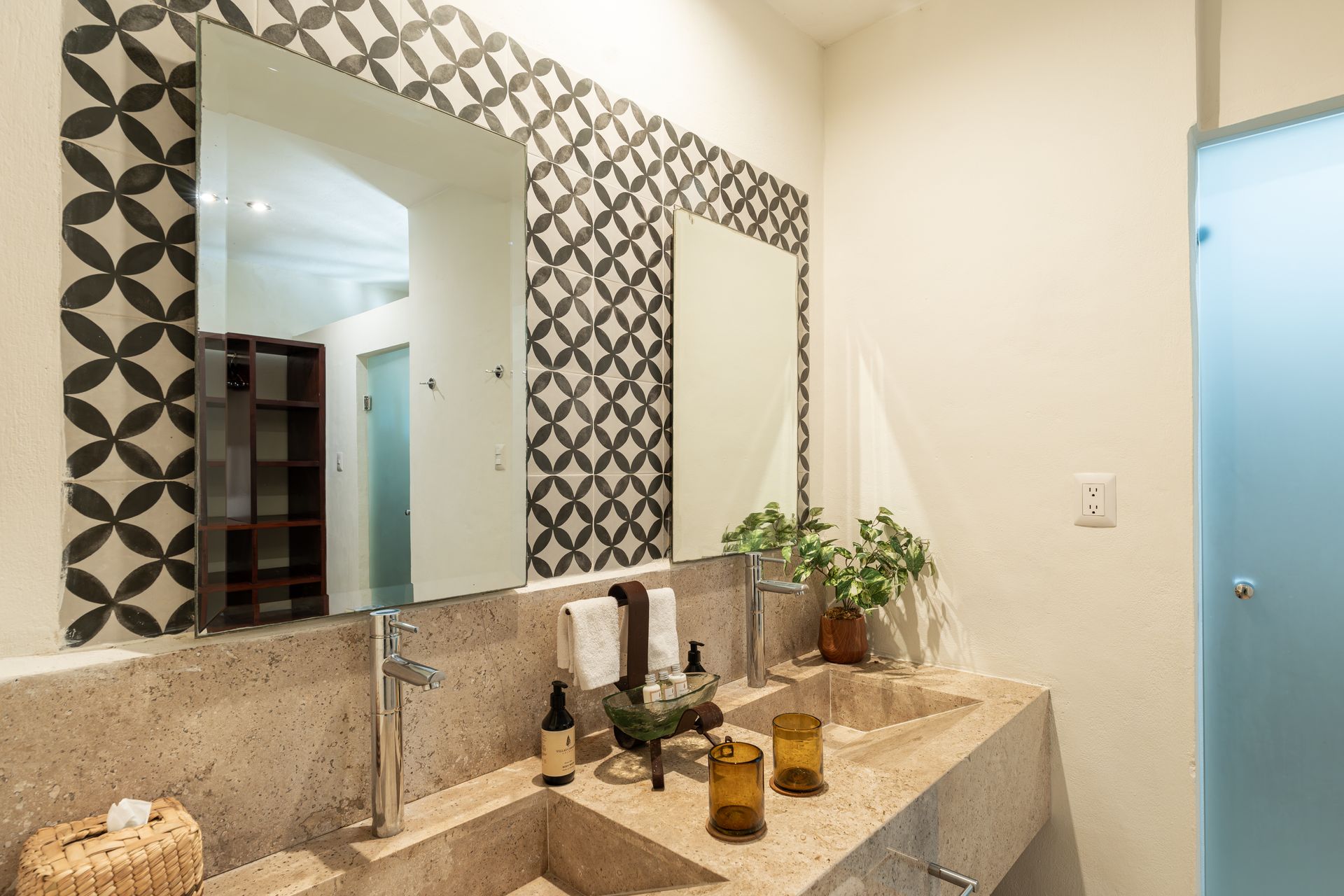 Bathroom with double sink, patterned mirror frame, beige countertop, potted plant, and frosted glass shower.