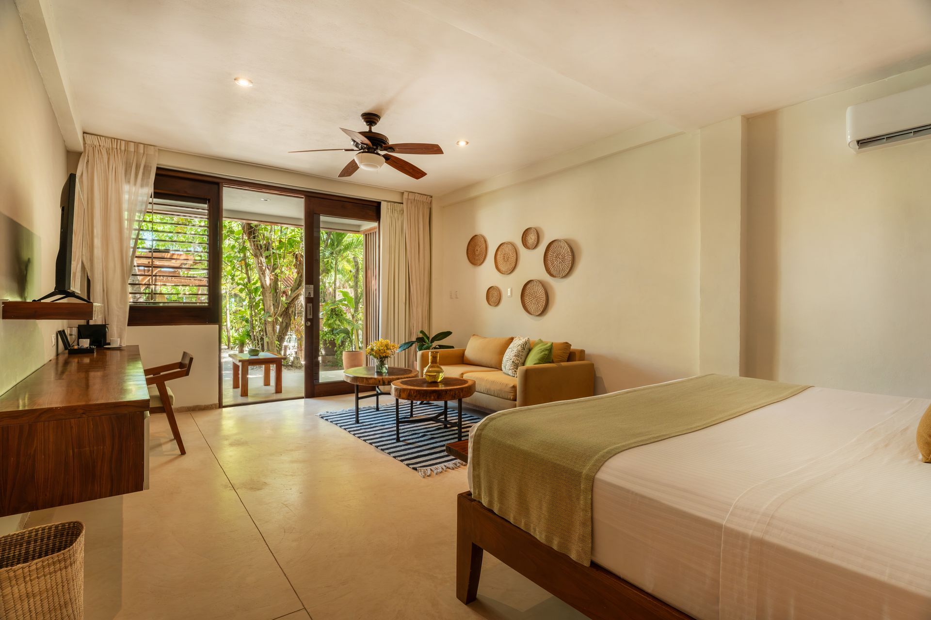 Bright hotel room with a bed, small sofa, wooden desk, and a balcony overlooking greenery.