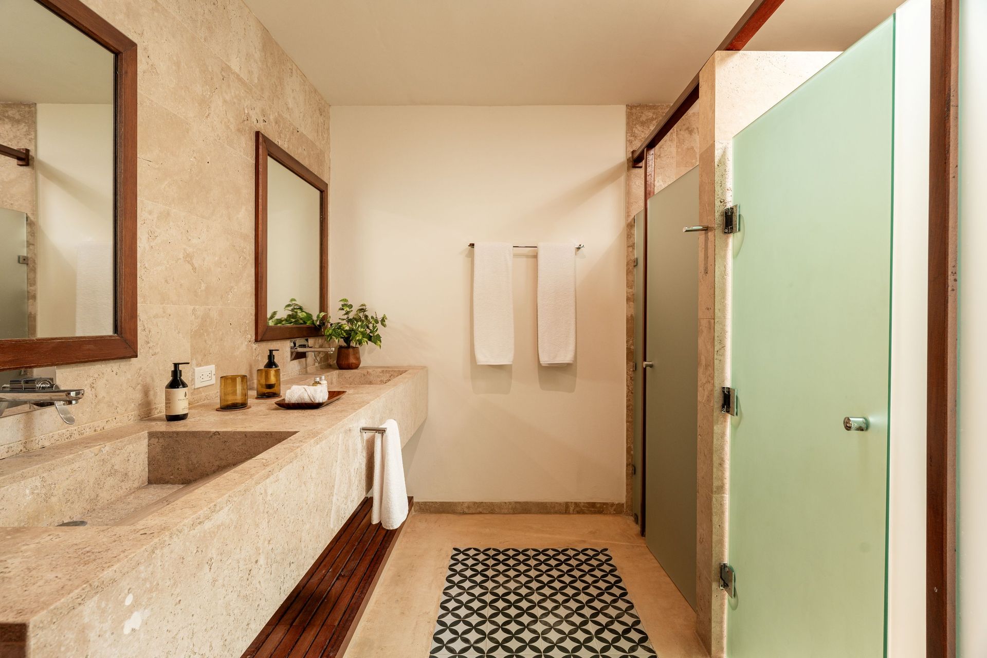 Modern bathroom with stone walls, double vanity, and glass shower door. Black and white patterned rug.