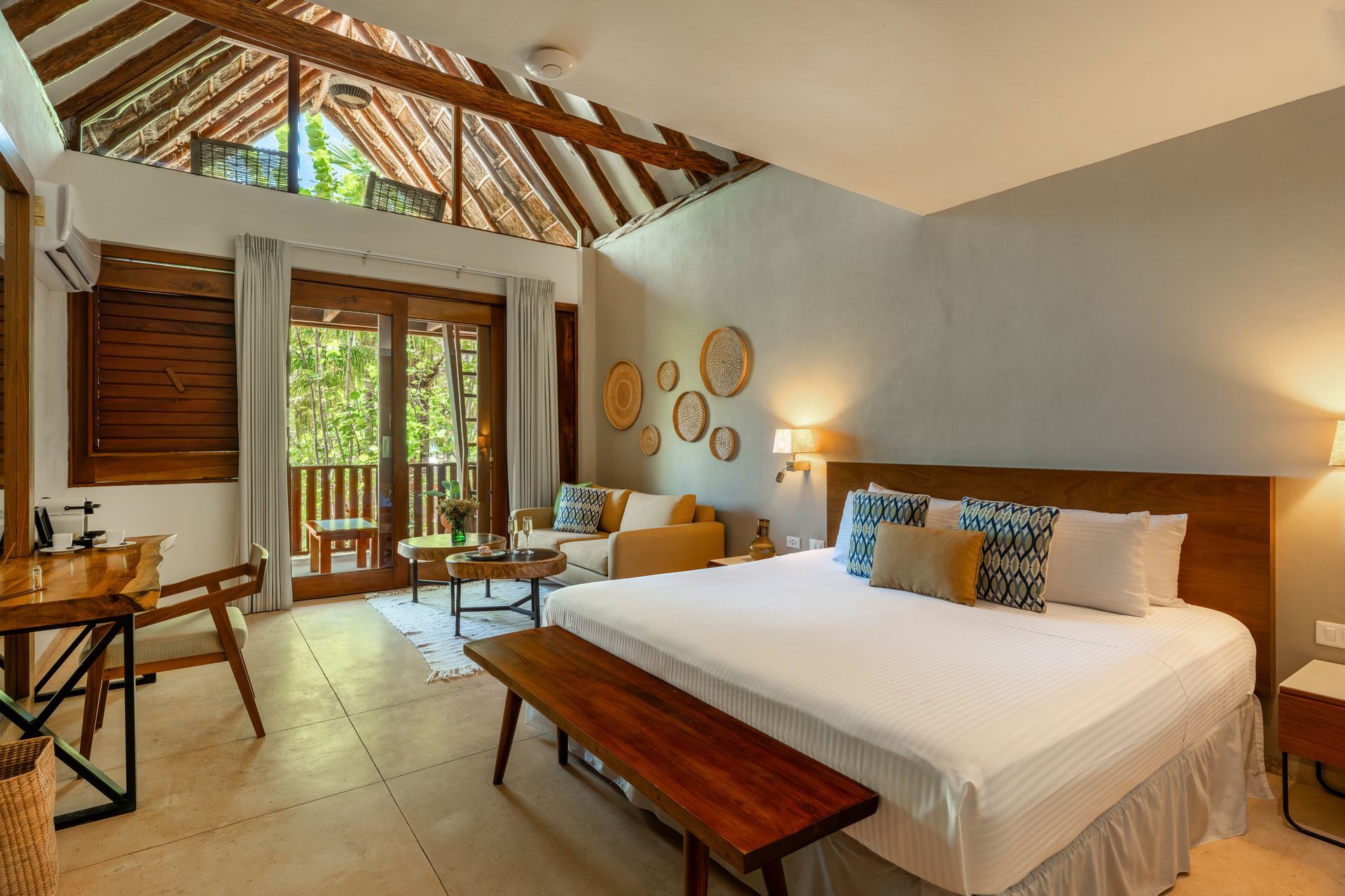 Bedroom with high ceiling, large bed, sitting area, wooden accents, and a balcony with greenery.