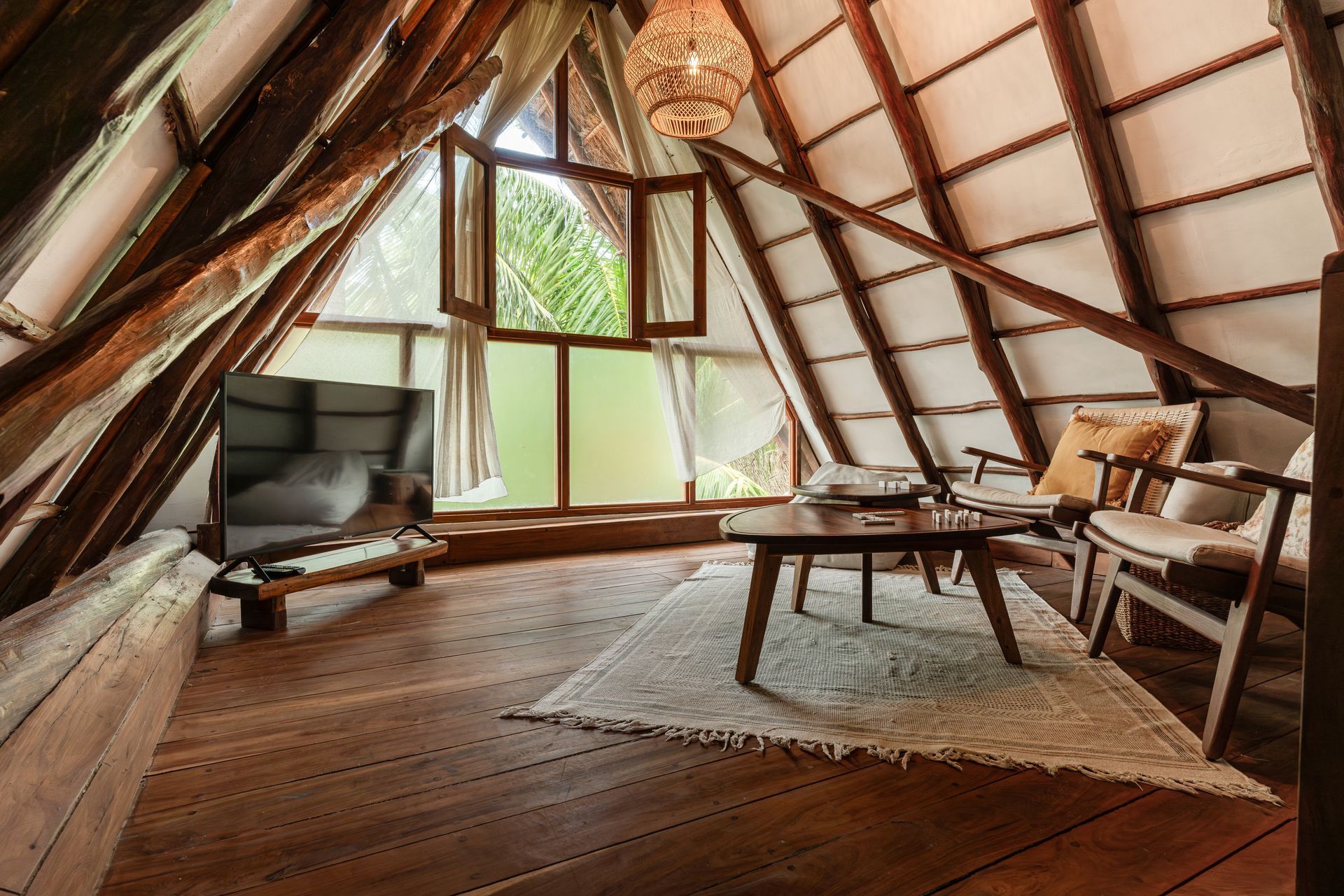 Cozy attic room with wooden beams, a TV, chairs, coffee table, and a window with a view.