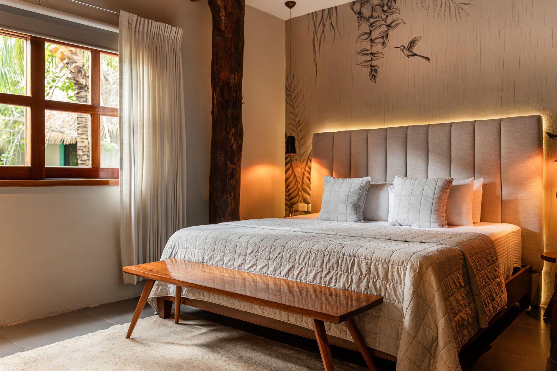 Bedroom with a king-sized bed, wooden bench, window with curtains, and decorative wall art. Warm lighting.