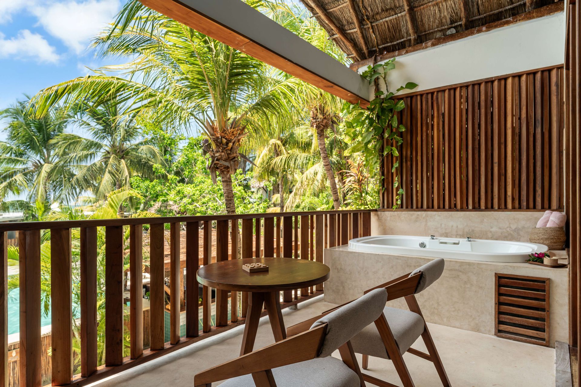 Balcony with table, chairs, and jacuzzi overlooking tropical trees under a blue sky.