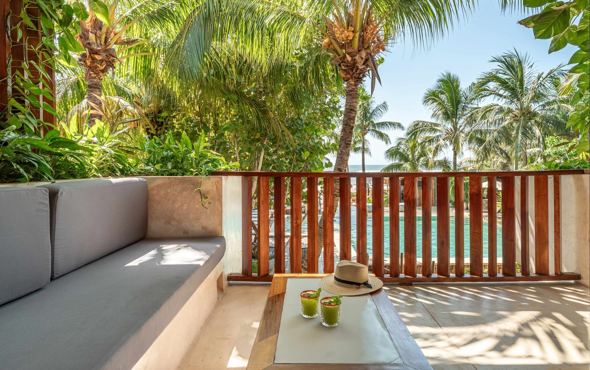Balcony with a built-in couch, coffee table, and drinks, overlooking a pool and palm trees.