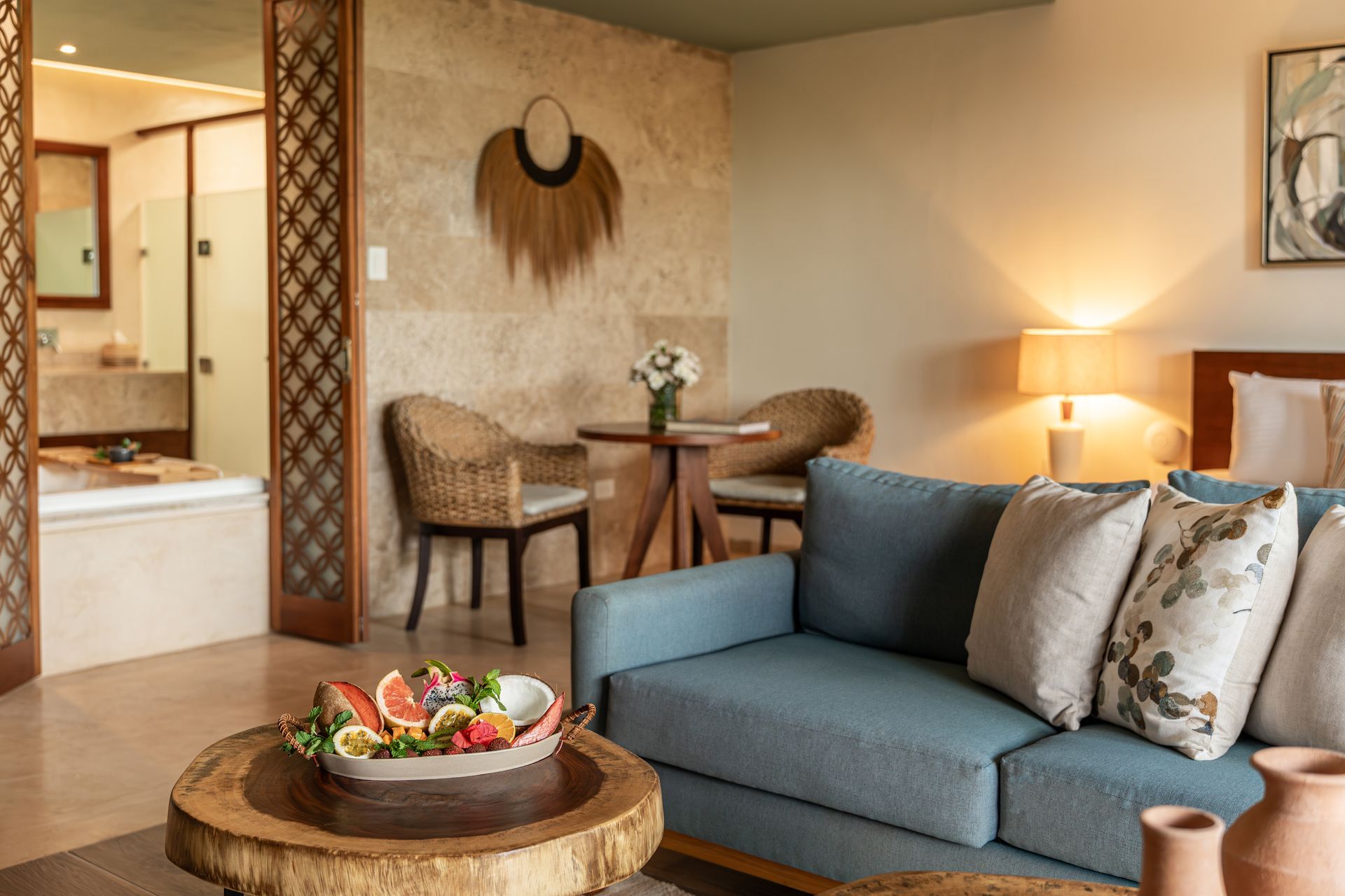 Cozy hotel room with blue sofa, wooden coffee table with fruit, and bathroom visible.