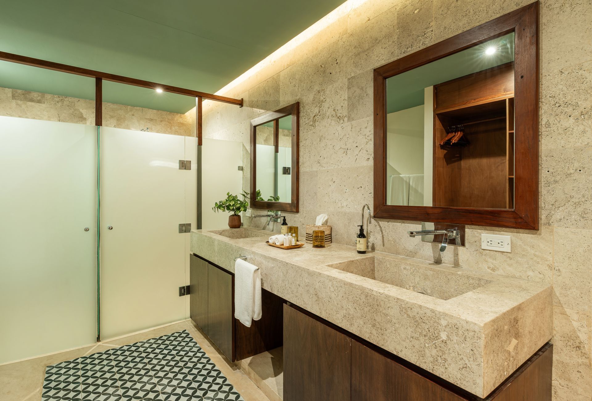 Modern bathroom with double sinks, frosted glass shower, wooden cabinetry, and tan speckled countertop.