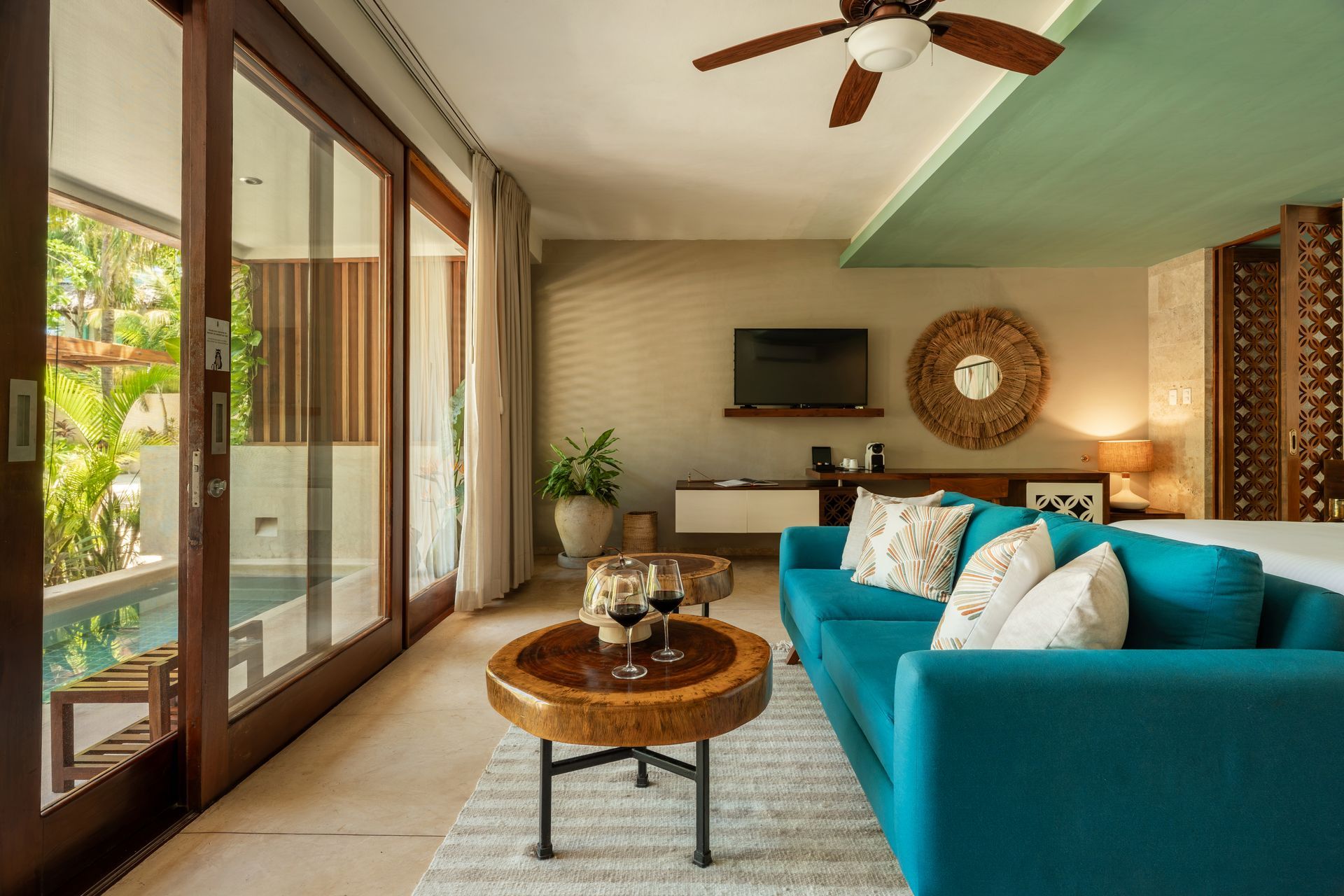 Cozy teal-accented living space with a pool view, featuring a sofa, coffee table with wine glasses, and a ceiling fan.