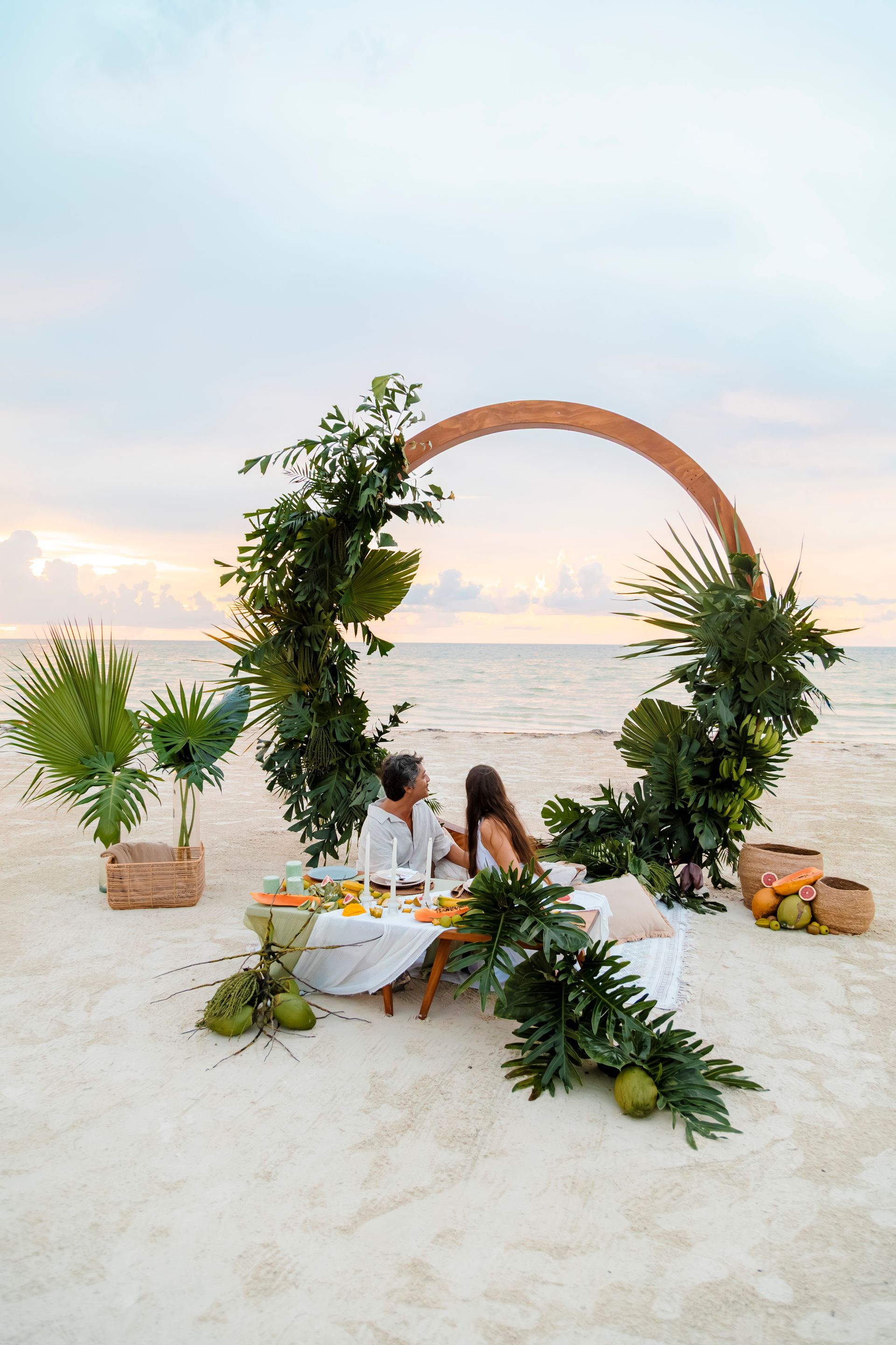 Couple at a beach dinner under a floral arch. Table with food; sand and ocean background.