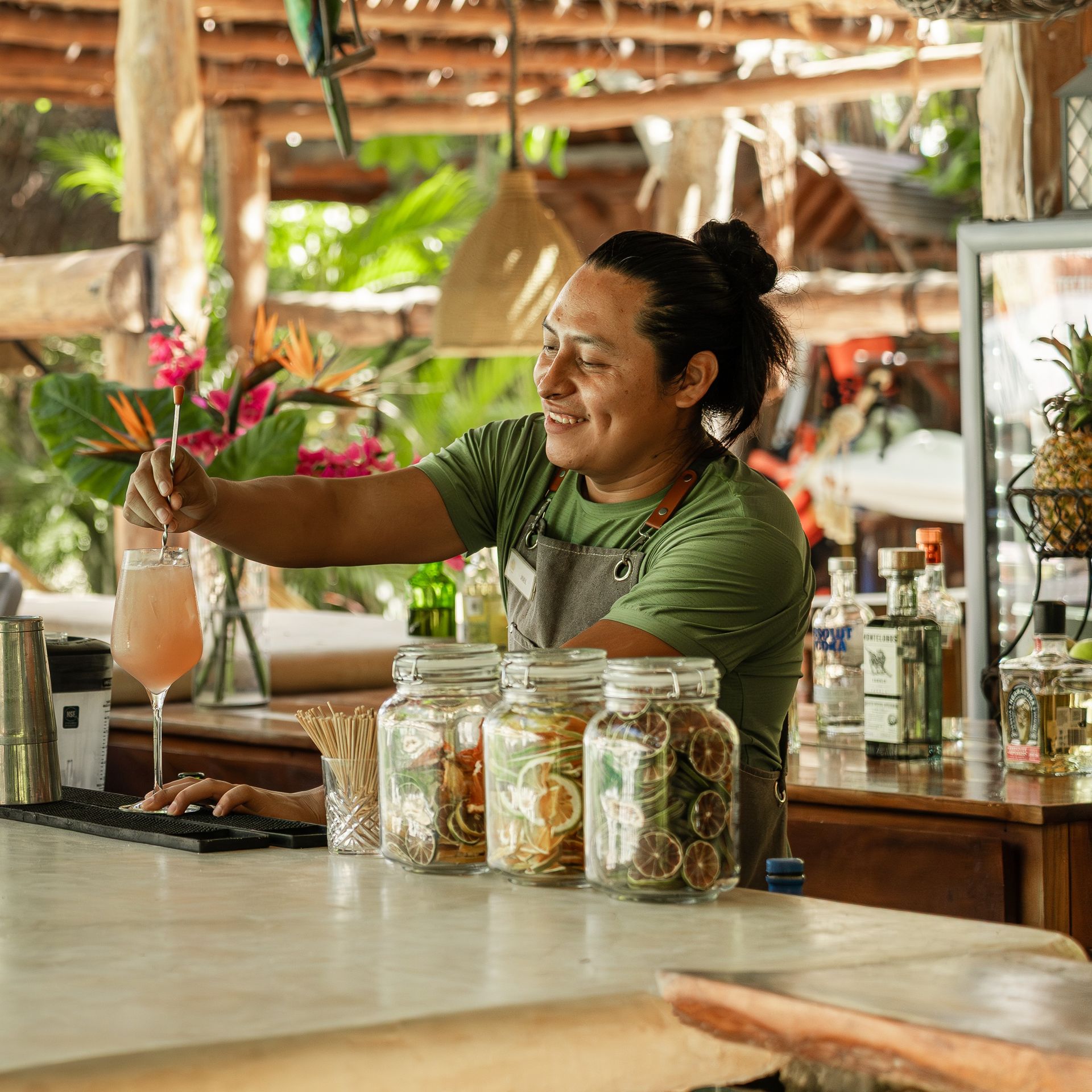 Bartender mixing drink at outdoor bar, smiling, surrounded by citrus garnish jars and bottles.