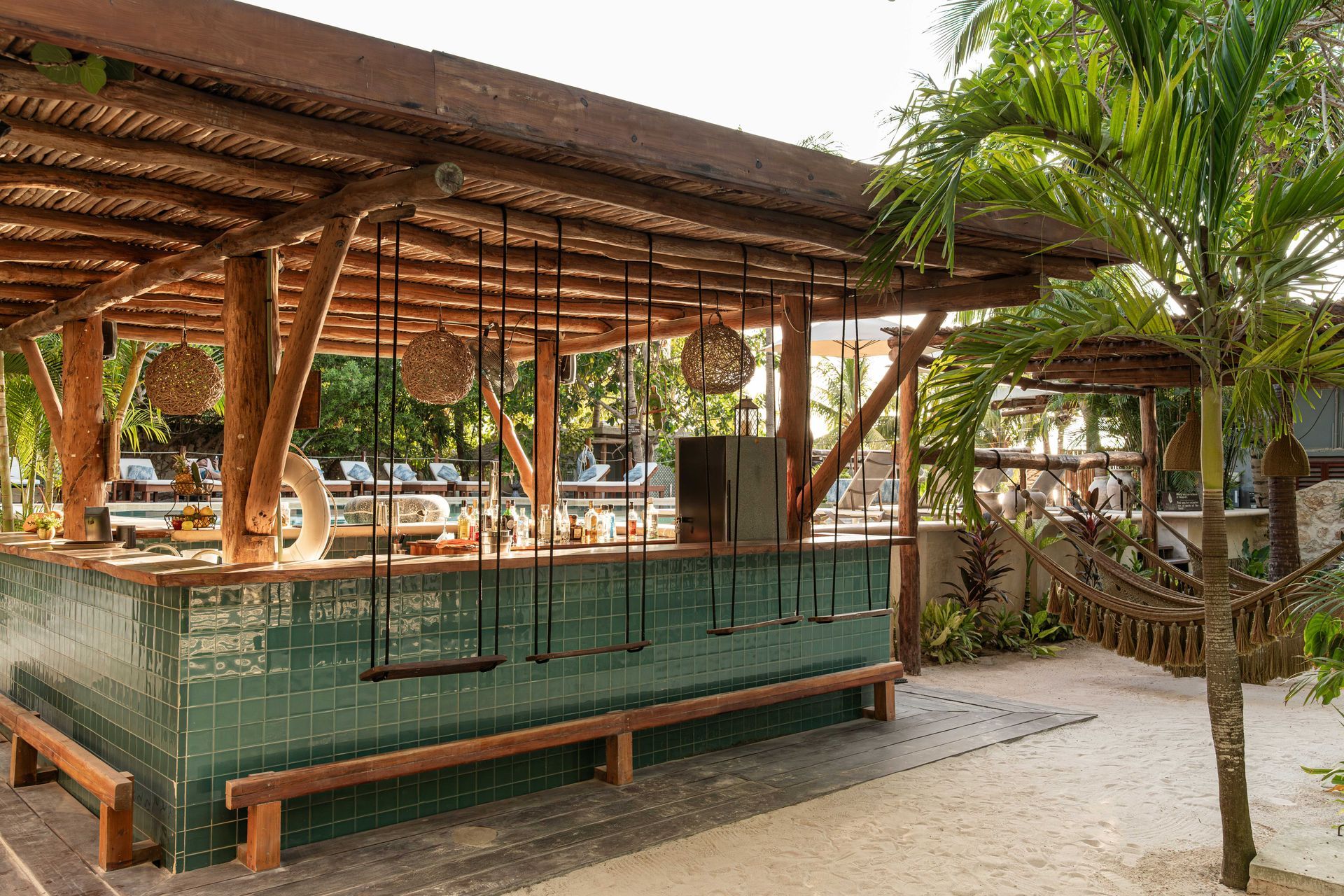 Outdoor bar with swings, thatched roof, and turquoise tiled facade. Sand ground, palm tree, and wooden benches.