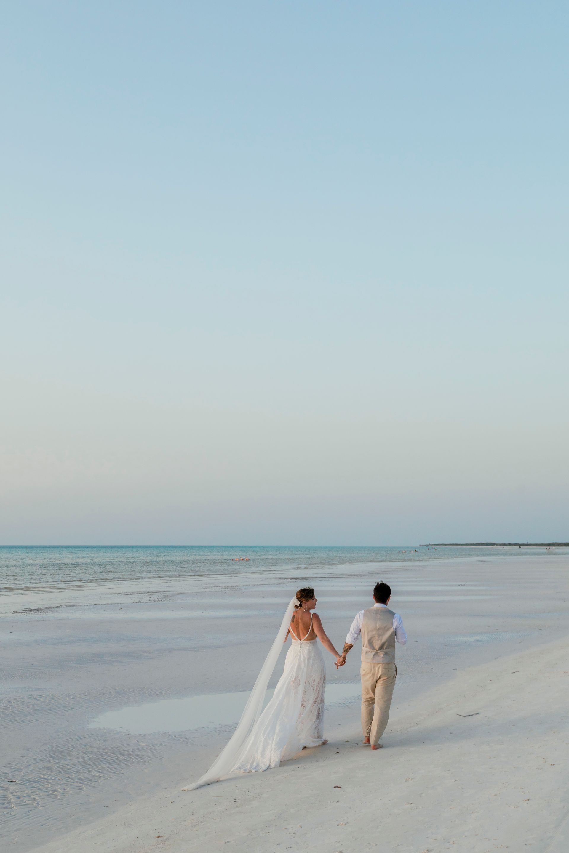 Couple holding hands walking on a beach at sunset; bride wearing a veil and dress, groom wearing beige pants.