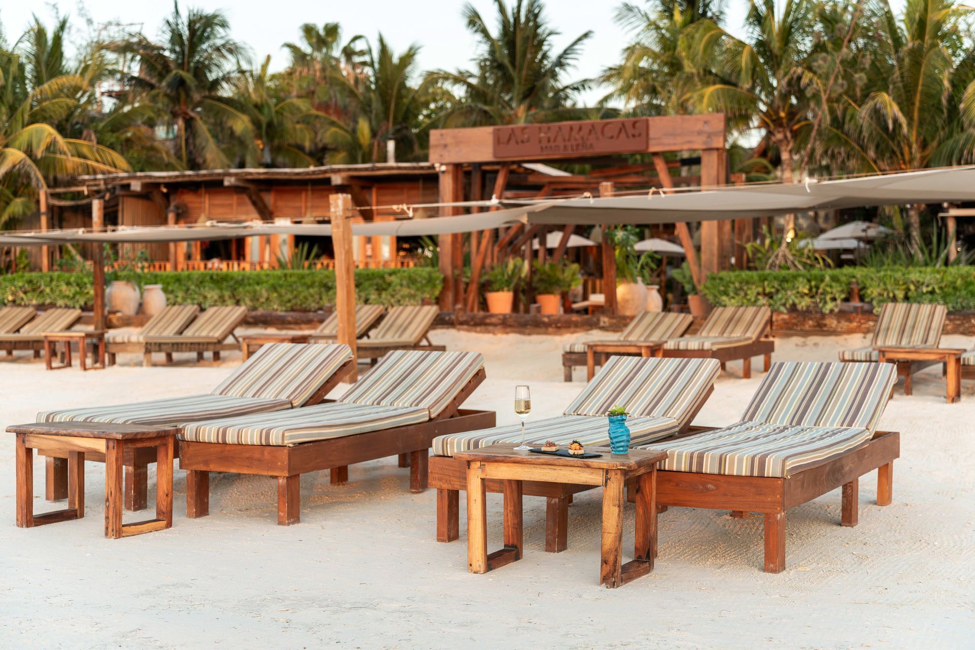 Beach chairs with striped cushions on a sandy beach, shaded by umbrellas, with a wooden structure in the background.