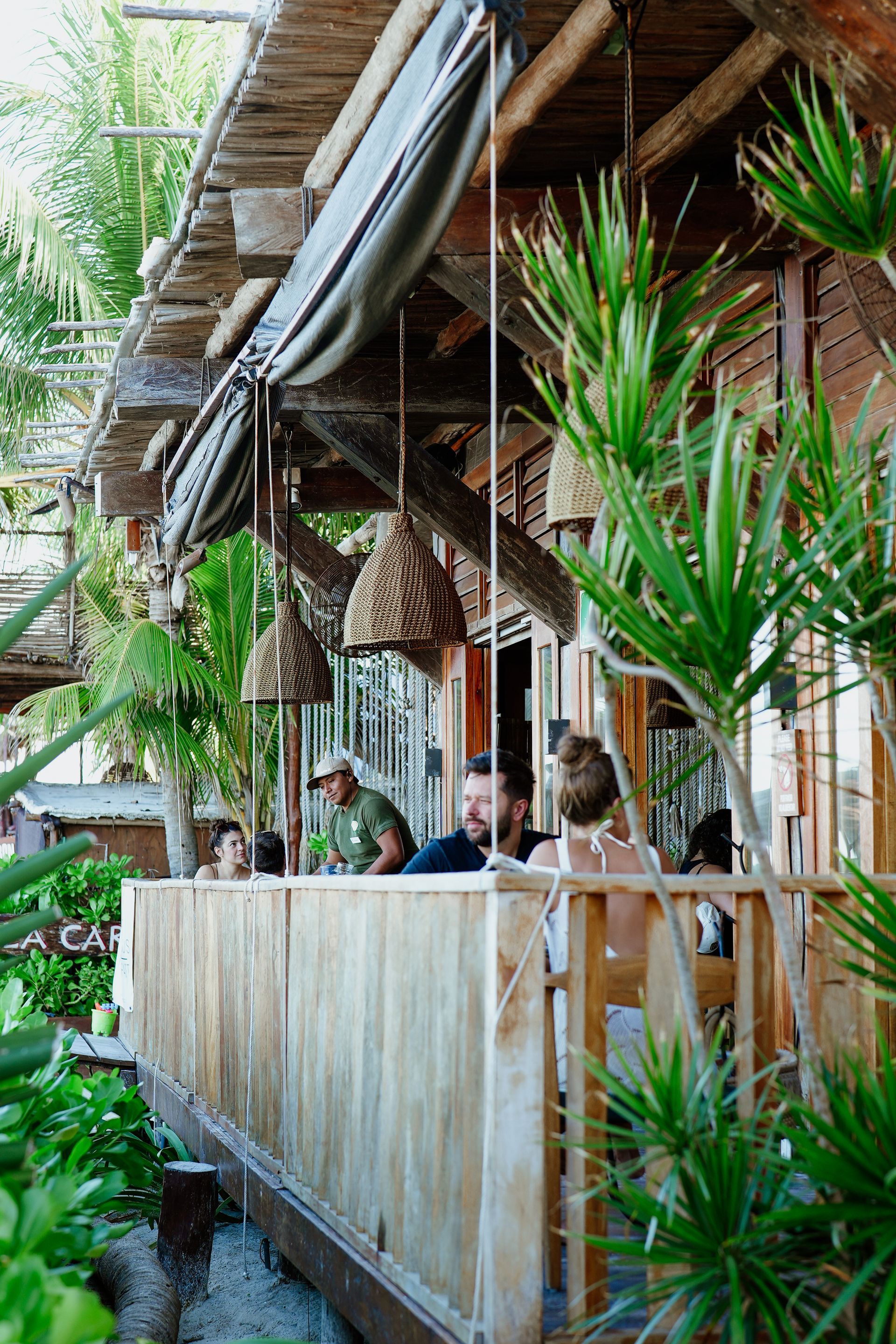Outdoor restaurant with wooden structure, hanging woven lights, and people dining. Green plants surround the patio.