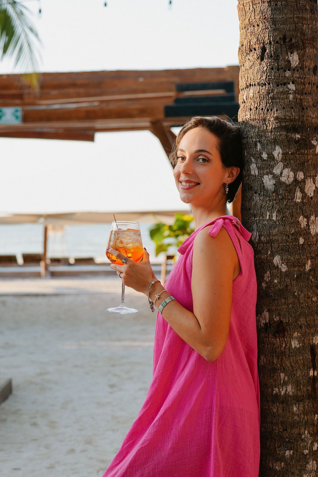Woman in pink dress holds an orange drink, leaning against a tree, smiling near the beach.