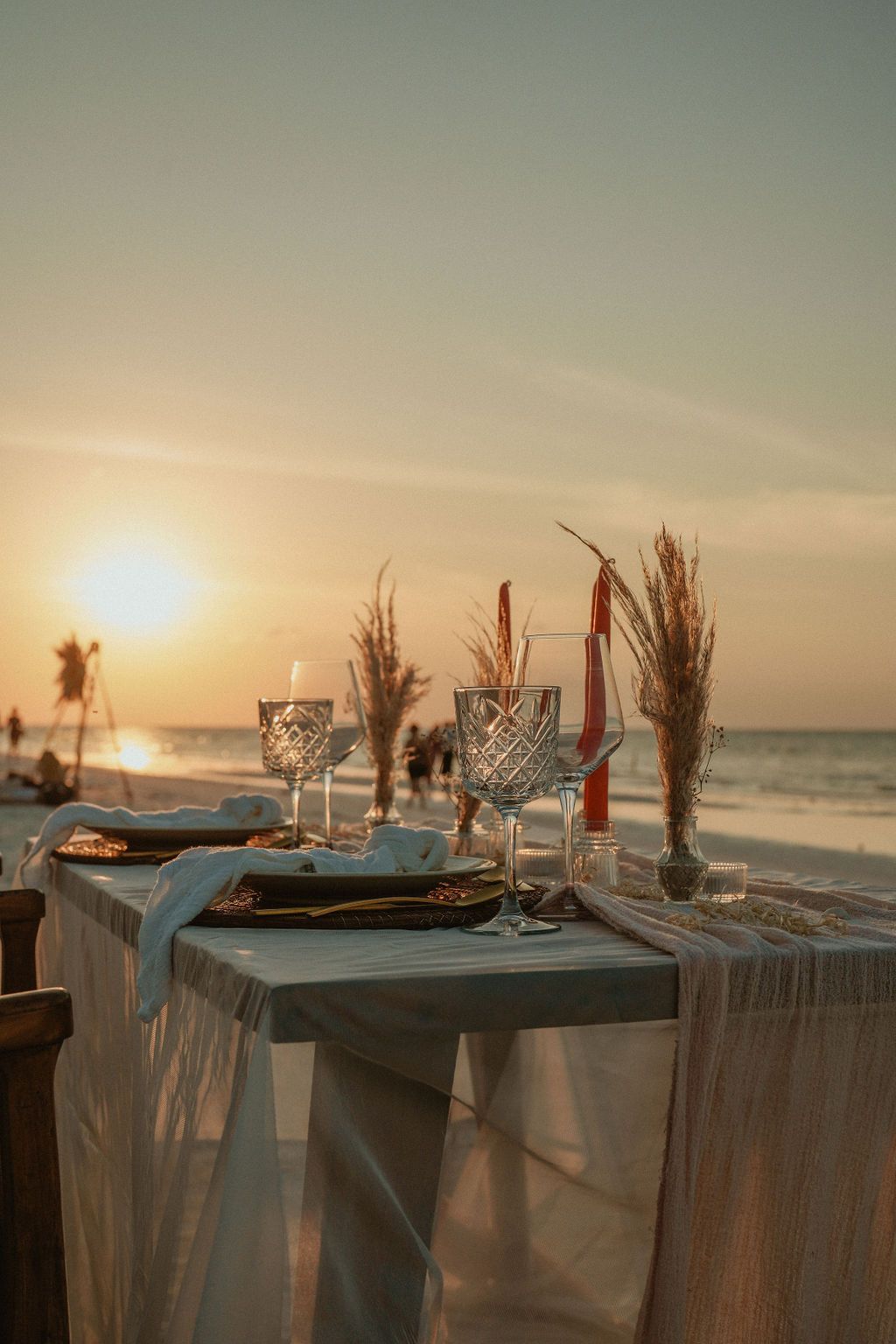Beach table setting at sunset with plates, glasses, and flowers.