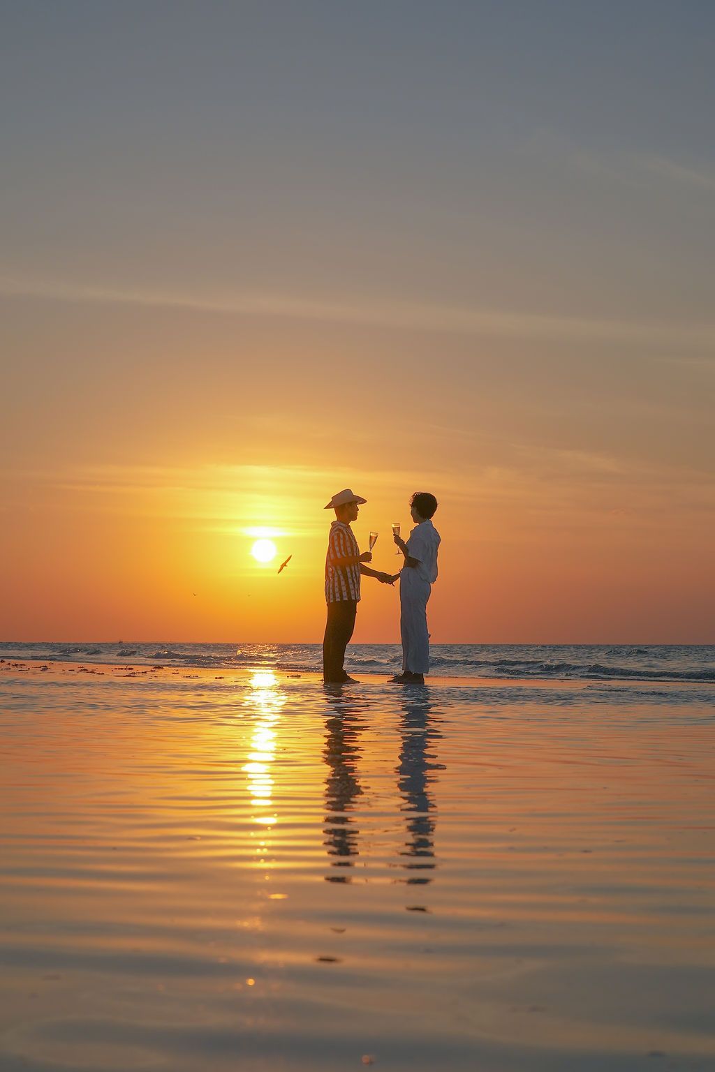 Two people stand on a wet beach, silhouetted against a setting sun reflecting on the water.