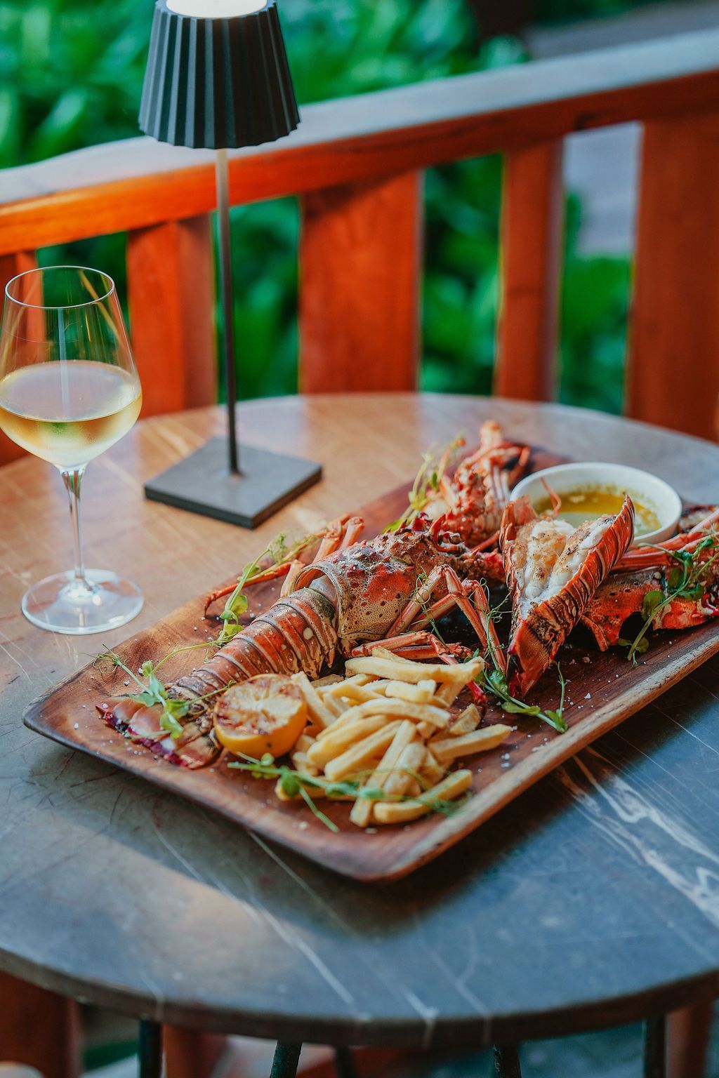 Lobster dish with fries and white wine on a wooden tray, table with lamp on a balcony.
