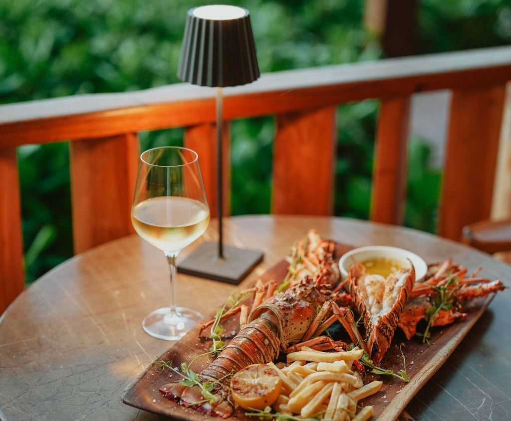 Grilled lobster and fries with wine, lit by a small lamp, on a wood table, balcony setting.