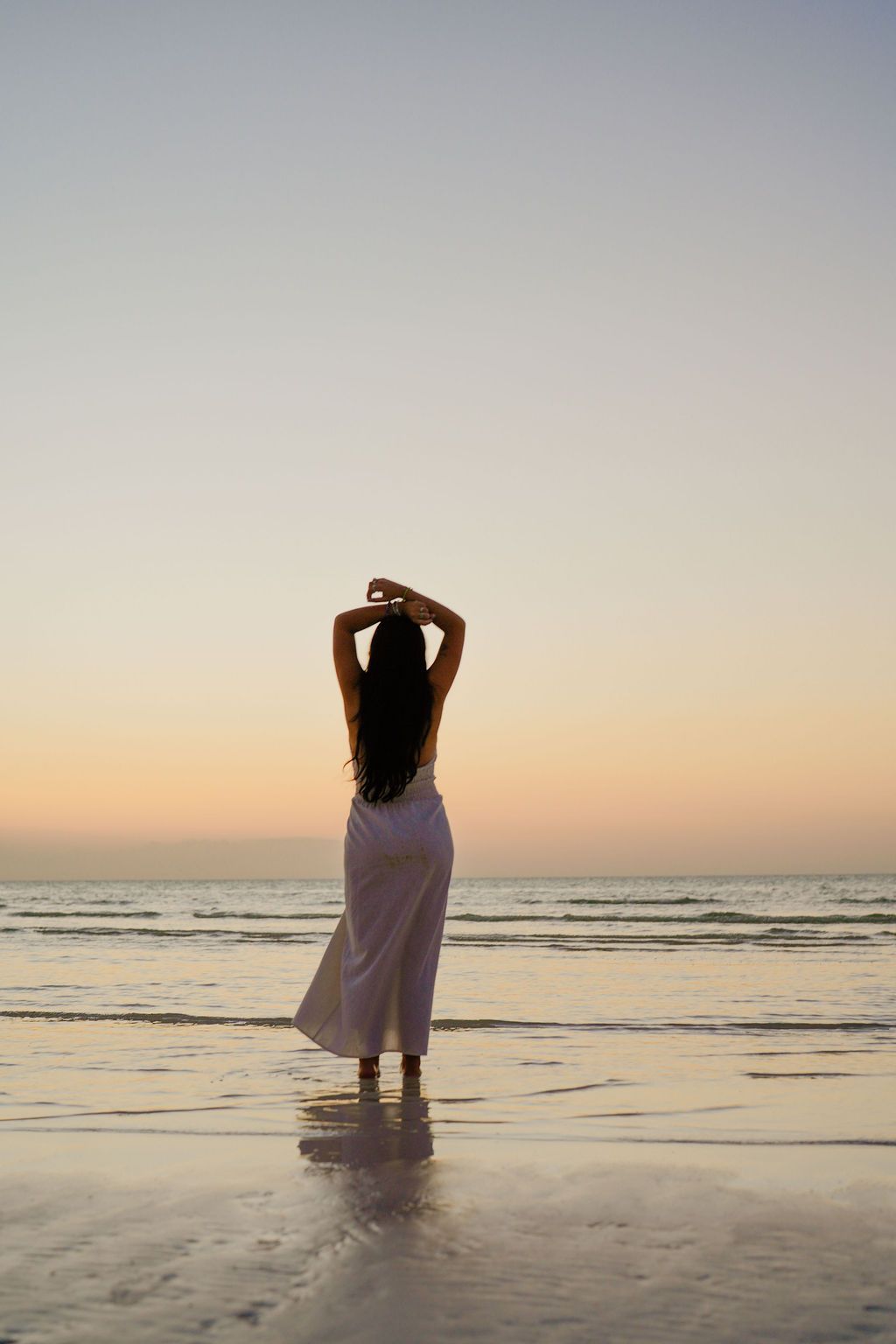 Woman in white dress with arms raised on beach at sunset.