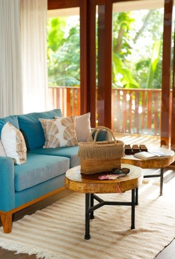 Blue couch, round wooden tables, and a woven tote bag in a bright, tropical-themed living room with open doors to a balcony.