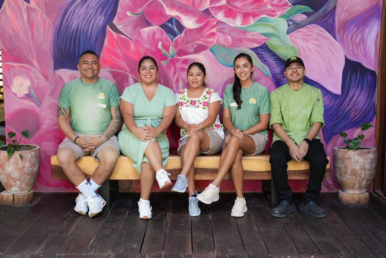 Five people in green shirts sit on a wooden bench against a floral mural.