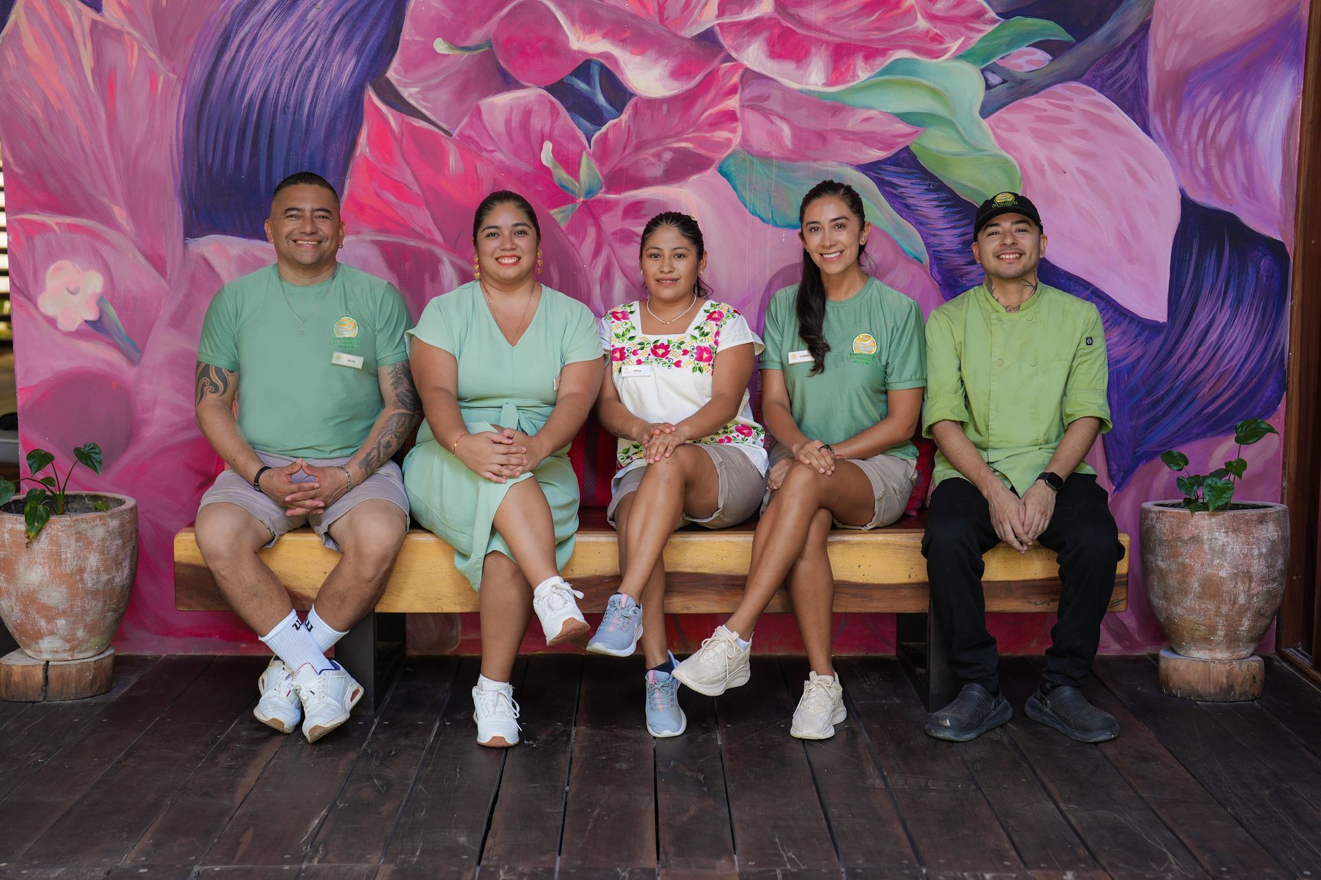 Group of five people in various shades of green, smiling, posing on a bench in front of a colorful floral mural.