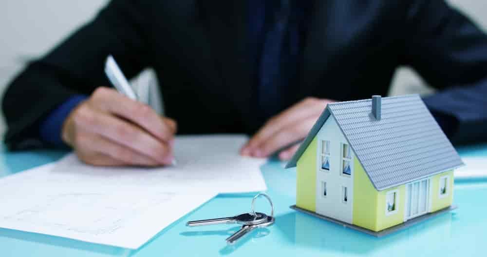 A Man is Writing on a Piece of Paper Next to a Model House and Keys — North Coast Conveyancing In Wauchope, NSW