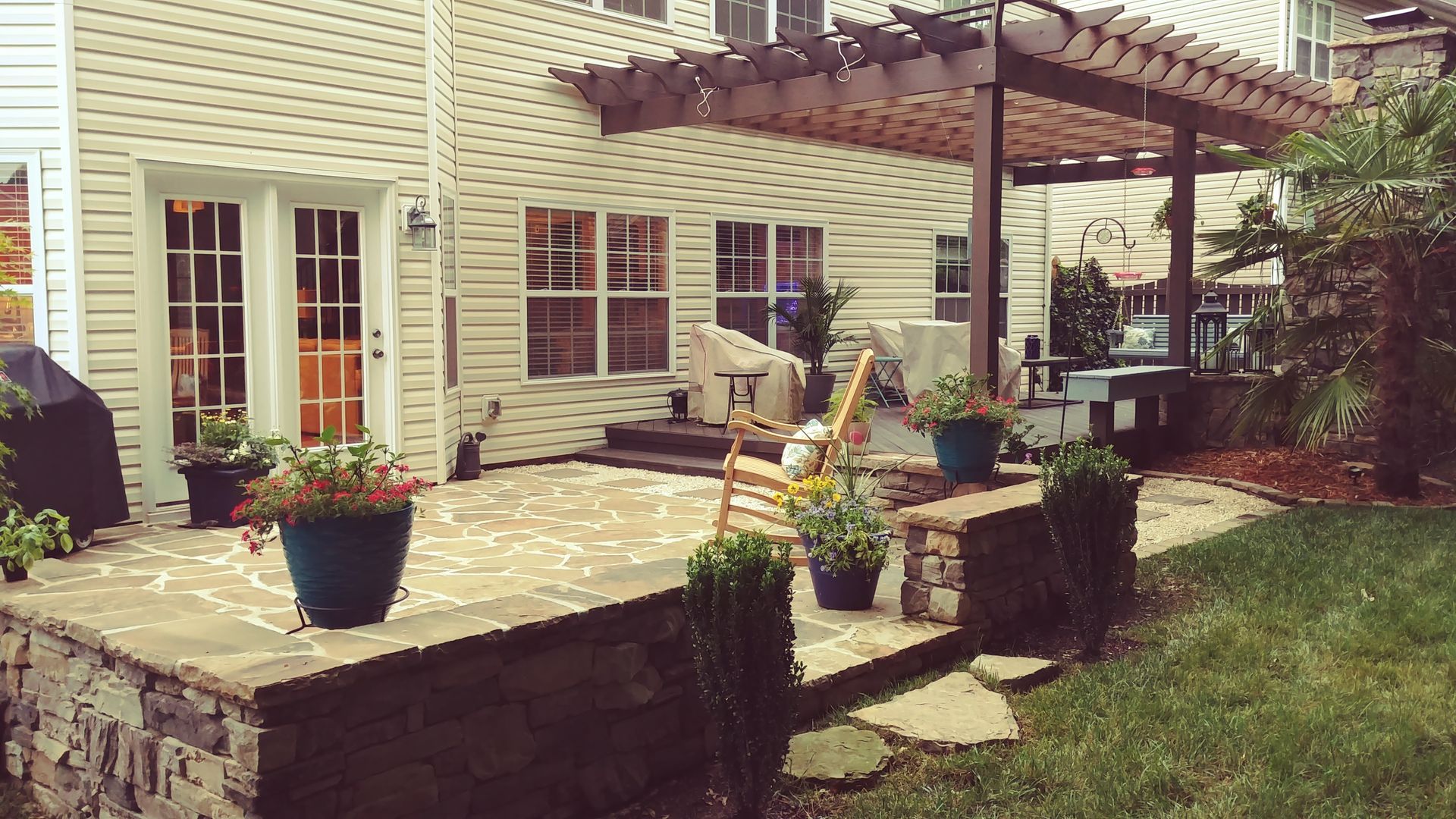 Patio with stone wall and pergola, potted plants, and backyard with house.