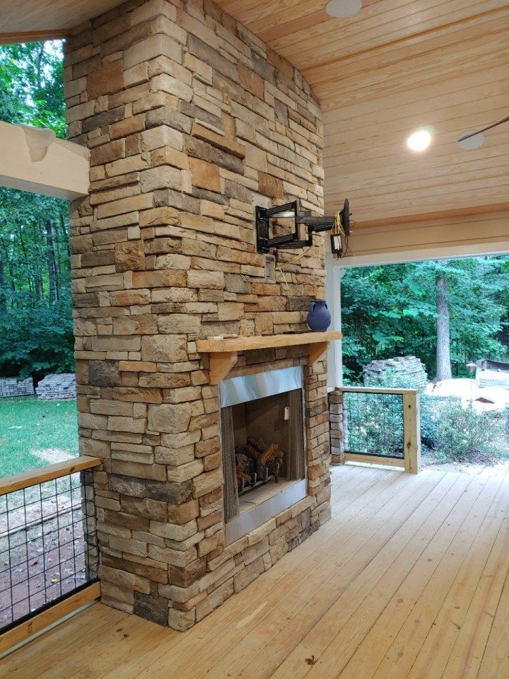 A stone fireplace with a wooden mantel on a covered outdoor deck with wood paneling and a view of trees.