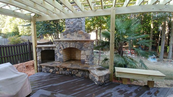 Stone fireplace under a wooden pergola on a wooden deck. Backyard setting with trees.