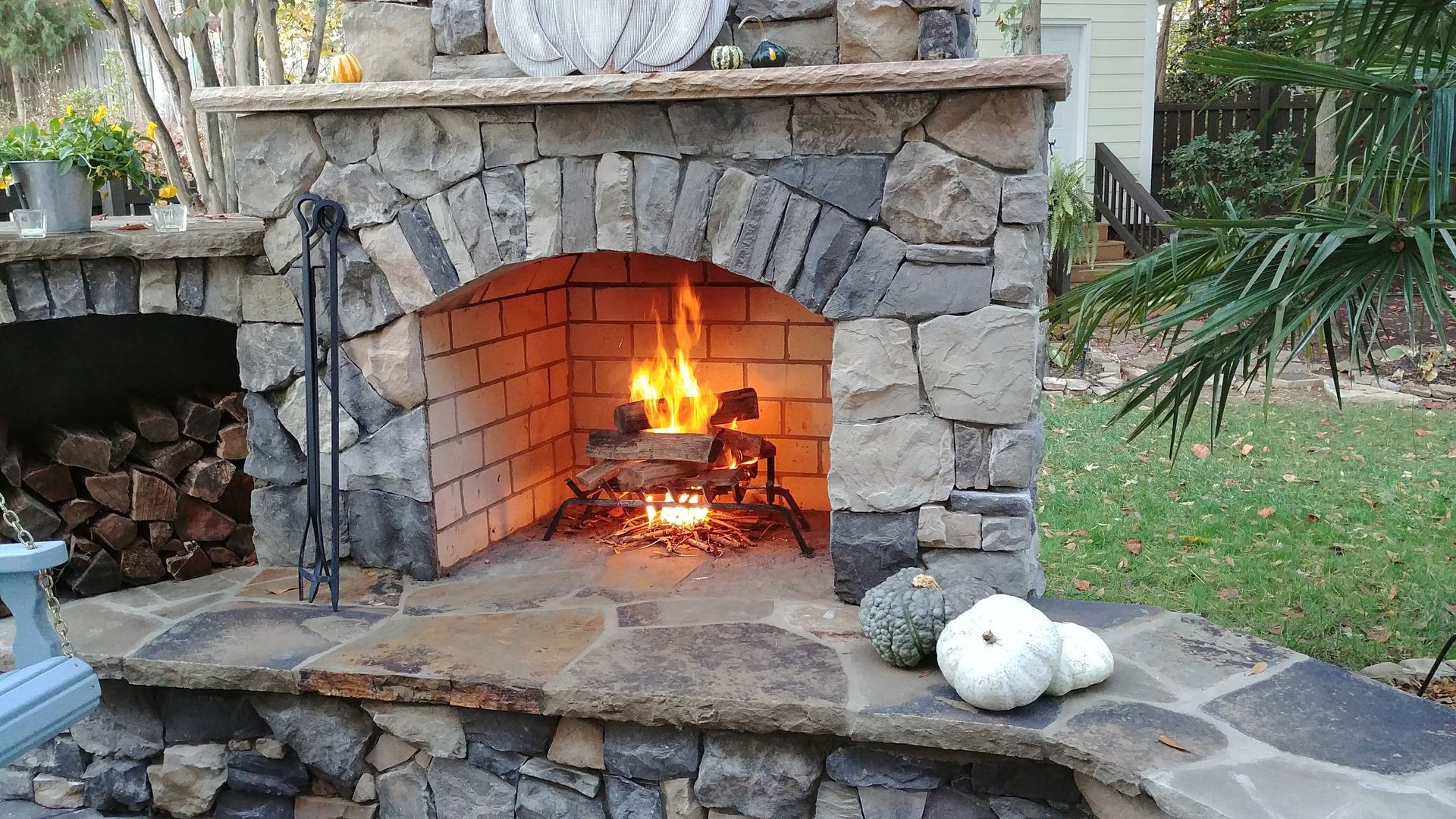 Outdoor stone fireplace with burning fire and stacked wood, pumpkins, and a green lawn.